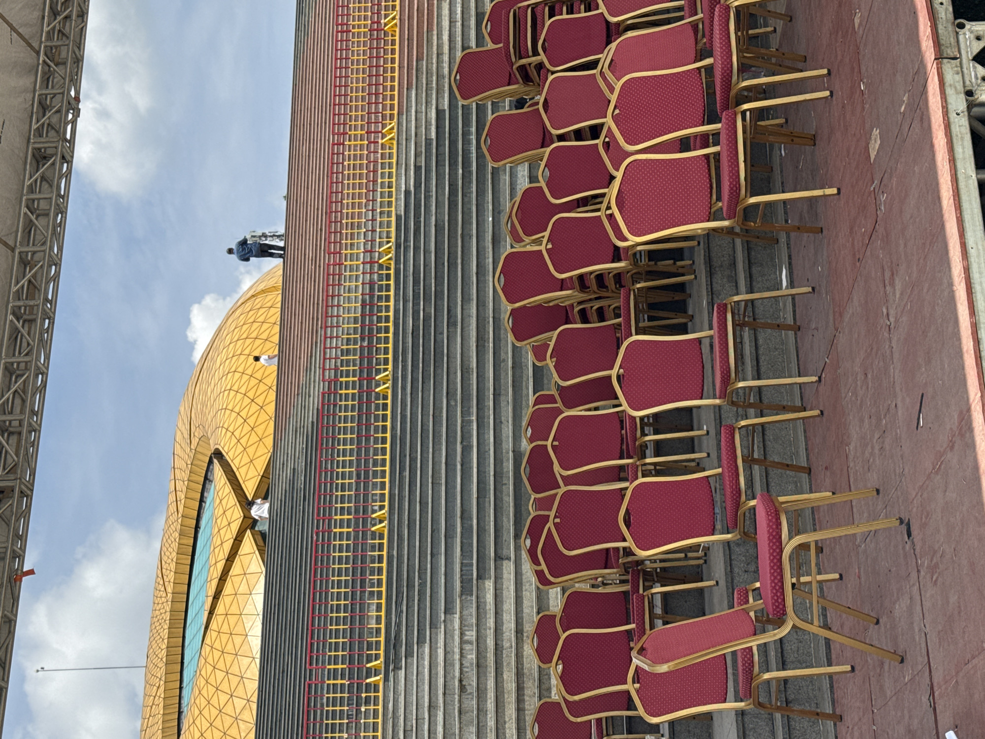 The image shows a view of a grandstand with rows of red upholstered chairs with gold frames. The chairs are arranged on a flat surface in the foreground, with some stacked further back. Behind the chairs are a series of concrete steps that lead up to a yellow, dome-shaped structure, possibly a building or part of a stadium. A metal fence with red and yellow bars runs along the edge of the steps. Two individuals are visible on the steps, one standing near the top and another further down. The sky is cloudy with patches of blue. The location is identified as Da Lat, Vietnam, suggesting this is likely part of an outdoor venue or event space in the city.