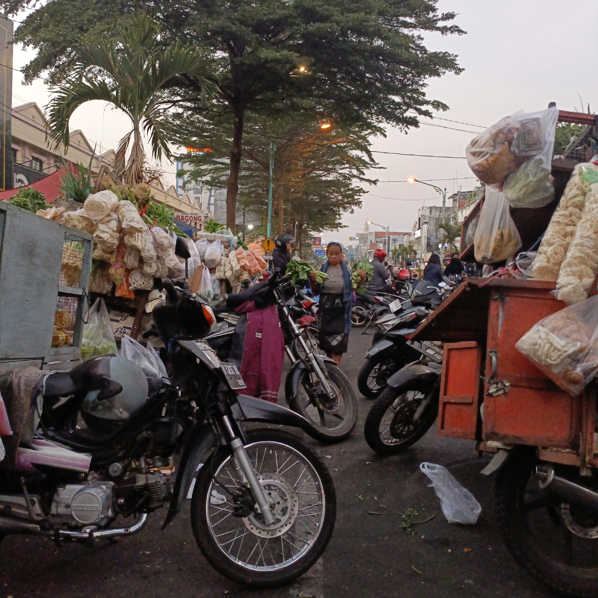 The image depicts a bustling street market scene in Salatiga, Indonesia, likely in the morning or late afternoon given the soft lighting and overcast sky. The foreground is dominated by several parked motorcycles, with their front wheels and engines prominently displayed. One motorcycle in particular, a dark color with a red and black seat, has its front wheel taking up a significant portion of the lower frame, showcasing its intricate spoke design and disc brake. To the left of this motorcycle, a gray metal cabinet with mesh doors is visible, containing various packaged food items.
Behind the motorcycles, a vibrant display of goods is being sold from carts and stalls. Piles of white, puffy snacks, possibly crackers or chips, are arranged in large bunches and packaged in transparent plastic. There are also bags of produce, including leafy green vegetables, and other dry goods.
Several people are engaged in market activities. A woman in the center of the frame, wearing a dark skirt and a blue scarf, is holding a bunch of green vegetables. She appears to be a vendor or a customer. Another person, wearing a helmet, is visible near a motorcycle further back in the scene. In the background, more motorcycles and market stalls are visible, along with buildings and trees lining the street. The overall impression is one of a lively and active marketplace.