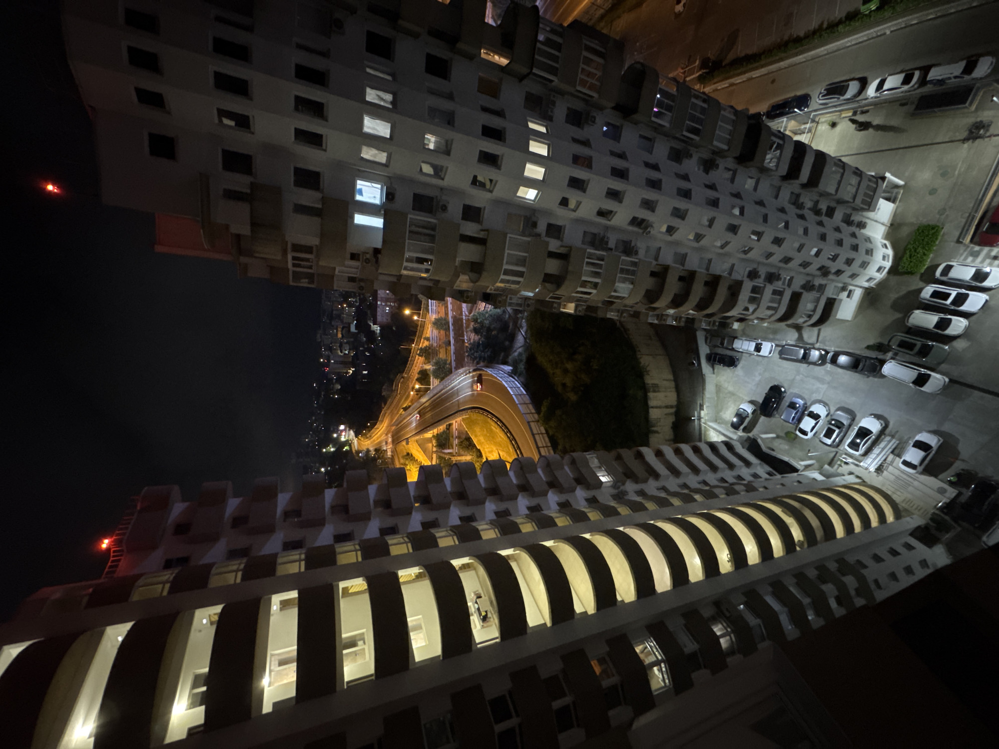 This high-angle, wide shot captures a view between two tall, modern apartment buildings at night in Sochi, Russia. The buildings are characterized by their balconies, which are stacked in a staggered pattern and illuminated from within. Many windows are lit, suggesting occupancy. A multi-lane highway is visible in the background, curving through the city and illuminated by streetlights. Cars are seen on the highway, and traffic lights can be observed. The surrounding urban landscape is dotted with more lights, indicating a densely populated area. Below the buildings, a parking lot is visible with several cars parked. One building has a red flashing light on its roof. The overall scene depicts an urban nighttime environment with typical city infrastructure and residential buildings.