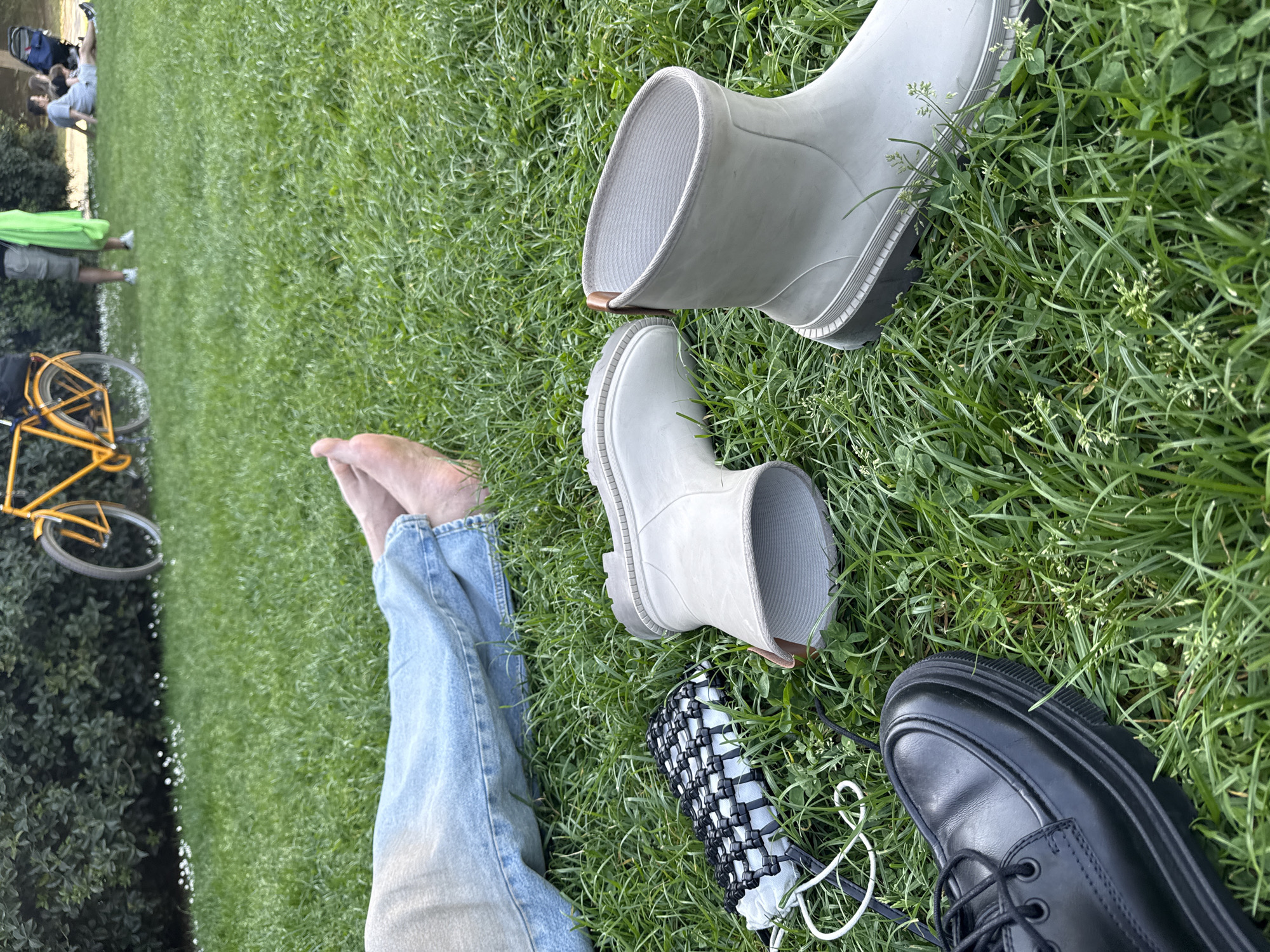 The image captures a high-angle view of a person relaxing on a vibrant green lawn, likely in a park. In the foreground, the person's bare feet and lower legs, clad in light blue denim jeans, are stretched out on the grass, suggesting a moment of rest and comfort.
Surrounding the feet are several items. To the right, two light grey/off-white rubber boots with thick, ridged soles are placed on the grass; one stands upright, while the other lies on its side. Further to the right, a single black lace-up shoe with a robust, lugged sole rests on the grass. A small, black and white woven bag, possibly a handbag, with white cord straps, is situated between the person's feet and the grey boots.
The setting is a natural grassy area, featuring various types of grass and some clover. In the mid-ground and background on the left side of the frame, elements of a public park environment are visible. A yellow bicycle with black tires is partially seen. Further back, several other people are present: one person wears a bright green top and dark shorts, standing, while a small group sits or lies on the grass with what appears to be a stroller or bag nearby, indicating a social and relaxed atmosphere.
The scene suggests a pleasant daytime outing in good weather. The person in the foreground has removed their footwear, emphasizing a sense of ease and relaxation. No visible text is present in the image. The overall environment aligns with a public green space or park, consistent with the provided context of Milan, Italy.