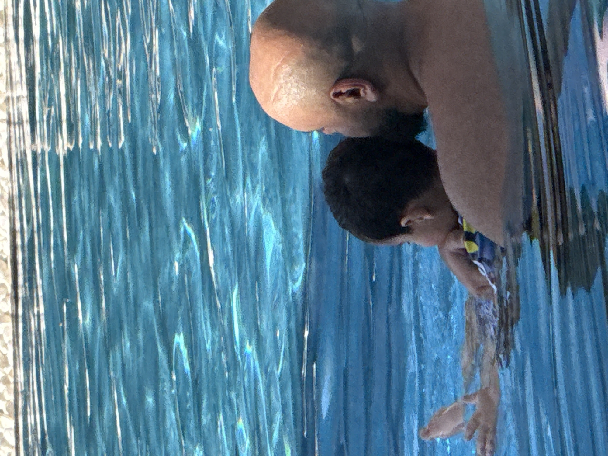 Two individuals are in a swimming pool. The person in the foreground is a bald adult male, facing away from the camera and looking towards the right. The person in the background is a child, also facing away from the camera, with their arm extended. The water in the pool is blue and reflects sunlight. The edge of the pool, made of small white stones, is visible at the top of the image. The scene depicts a recreational activity in a pool environment.