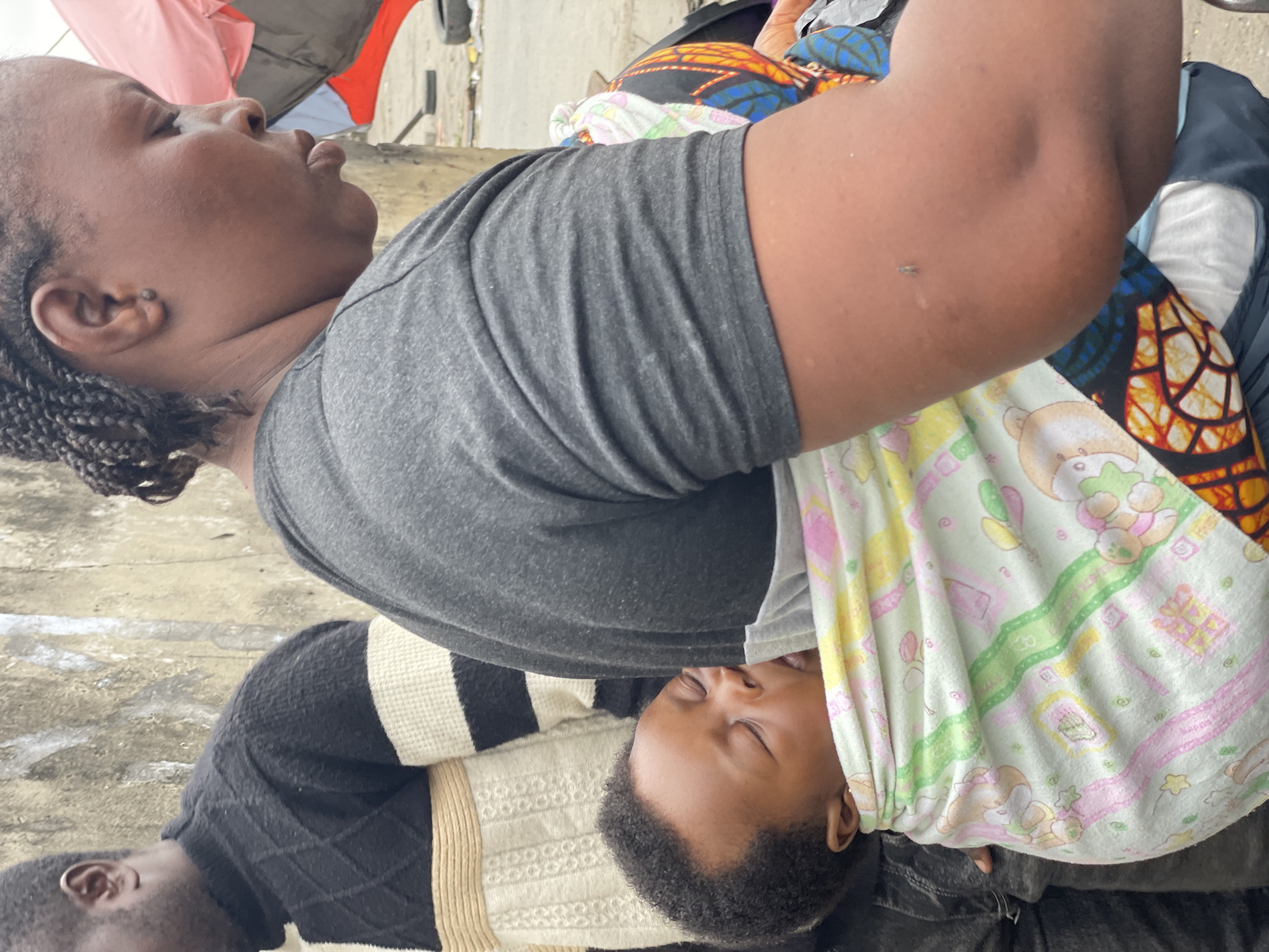 In Ado, Nigeria, a close-up, medium shot captures an African woman carrying a sleeping baby on her back using a patterned cloth. The baby, with eyes closed and mouth slightly open, is nestled against the woman's shoulder. The woman is wearing a grey t-shirt and her braided hair is visible. Beside her, partially visible, is a man wearing a black, white, and yellow striped sweater. The background suggests an outdoor urban environment with a textured wall and some debris on the ground.