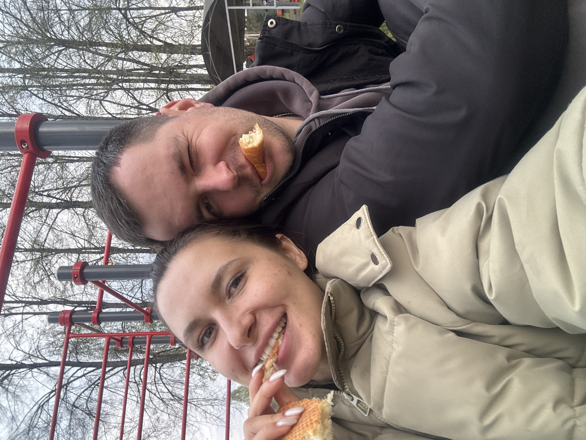 A selfie shows a man and woman eating outdoors in Красный Маяк, Russia. The woman smiles and holds a piece of bread to her mouth with her left hand. She wears a light-colored, puffy jacket. The man lies behind her, winking, with a piece of bread in his mouth. He wears a black jacket over a gray hooded sweatshirt. In the background are red metal poles, bare trees, and a structure.