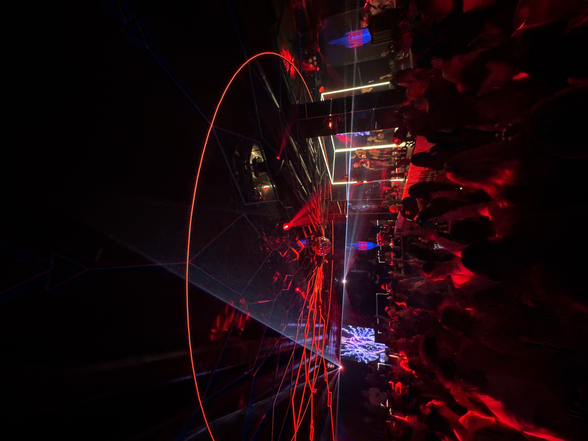 The image depicts an indoor nightclub or live performance venue located in Sofia, Bulgaria. The scene is characterized by dim ambient lighting and dynamic red laser projections, forming intricate patterns across a faceted or mirrored ceiling structure, which includes a prominent circular red laser ring. A disco ball is suspended from the ceiling.
A large, densely packed crowd occupies the foreground and midground, facing an elevated stage located towards the right side of the frame. On this stage, multiple performers are visible, appearing to be engaged in a live musical act, with at least one individual holding a microphone. The stage area is illuminated by vertical light strips. To the left of the stage, a large video screen displays abstract purple and blue fractal-like visual effects. The crowd, largely indistinct due to the low light and motion blur, is comprised of individuals standing, some with raised arms, indicating active engagement with the performance. Reflective surfaces on columns or walls near the stage amplify the visual effects of the environment.