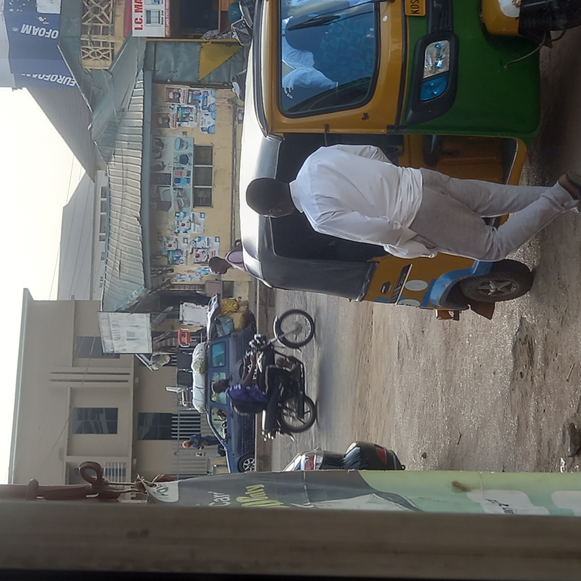 A man wearing a white, long-sleeved shirt and gray pants is standing on the passenger side of a yellow, green, and blue tricycle. The tricycle is on a paved road in Sardauna, Nigeria. A motorcycle and car are parked behind the tricycle. In the background are buildings with signs, including one for I.C. MA. and Eurofoam.