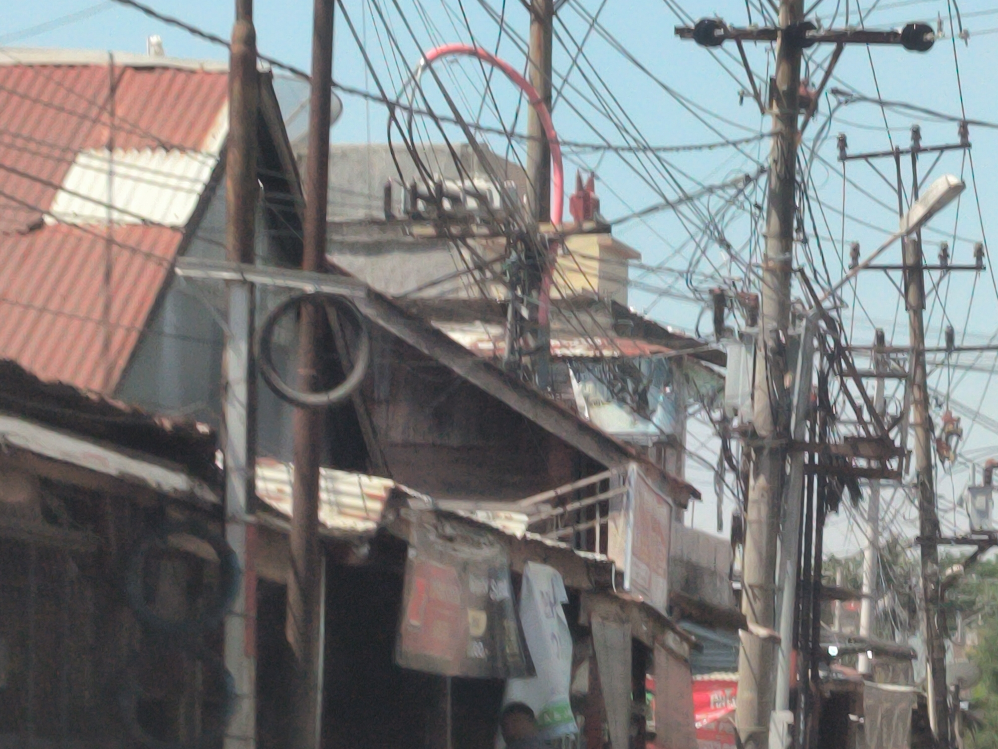 The image captures a densely populated urban scene in Lhokseumawe, Indonesia. A jumble of electrical wires crisscrosses the sky, connecting a series of utility poles. The structures below are a mix of weathered buildings, primarily one-story with corrugated metal roofs in shades of red and white. Several of the structures appear to be shops, with advertising signage visible, and garments hanging outside. A red arc-shaped object stands in the background on top of one of the buildings. The overall impression is one of a cluttered and perhaps economically challenged neighborhood. The weather appears to be clear and sunny.
