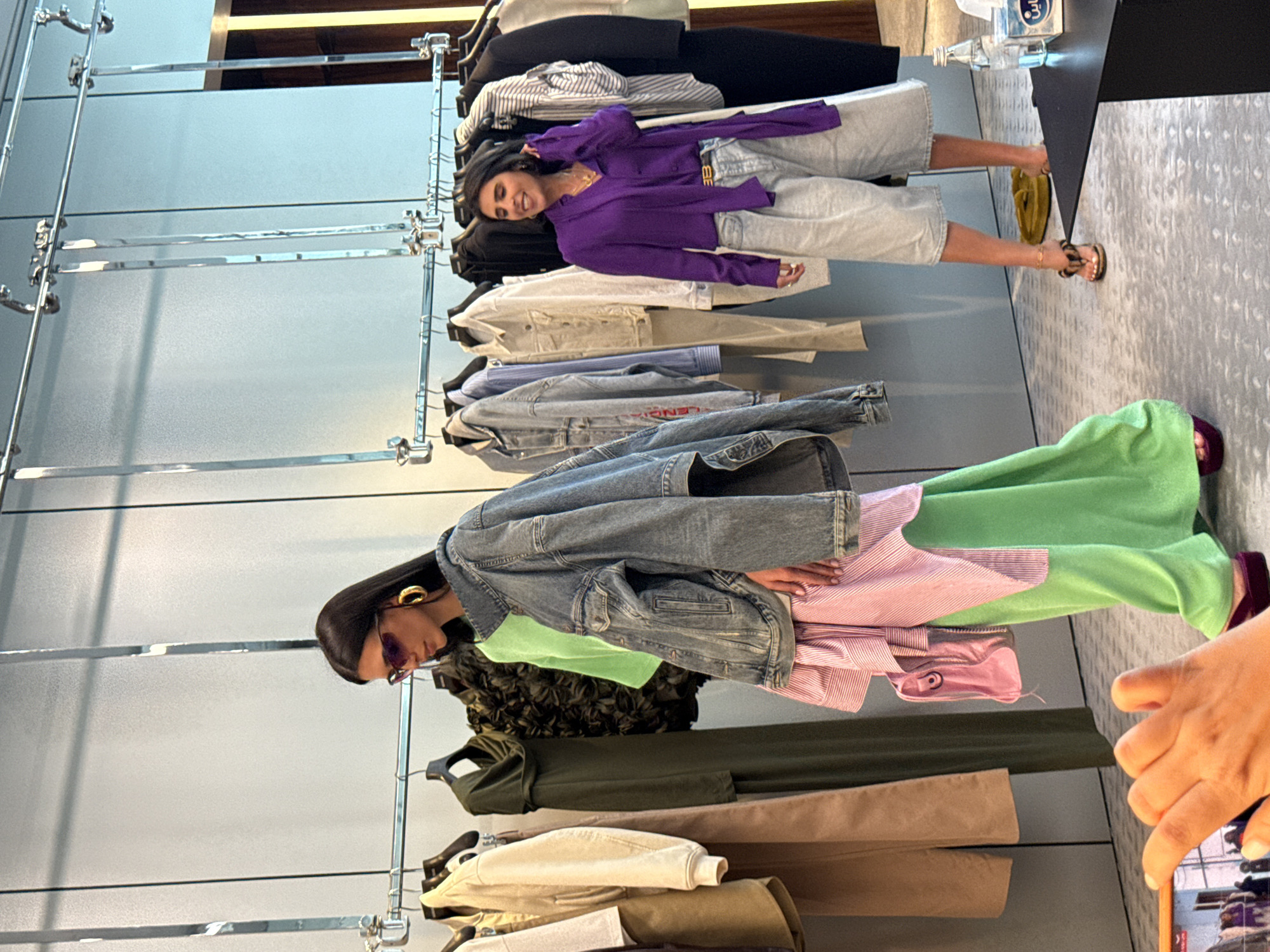 In a retail store in Dubai, United Arab Emirates, two women are observed. On the left, a woman with dark hair styled in a bob and wearing large gold hoop earrings and sunglasses, is dressed in a neon green dress layered with a pink and white striped shirt and a denim jacket. She is holding a small pink purse. On the right, another woman with dark hair is wearing a purple blouse with a scarf detail, paired with light-wash denim shorts and leopard print sandals. She is smiling and touching her hair. They are standing amidst racks of clothing displayed on chrome hanging rails. A black table with a bottle of water and a tissue box is visible in the foreground. The floor has a textured pattern. A hand holding a smartphone is in the lower left corner, capturing the scene.