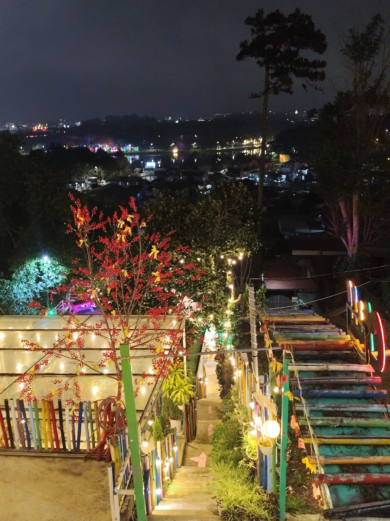 The image shows an outdoor staircase leading down a hillside in Da Lat, Vietnam, at night. The staircase is constructed of logs painted in various colors and is illuminated by string lights. A decorative tree with red berries and yellow ribbons stands to the left of the staircase. To the left of that is a wall made of painted stakes. In the background, the city lights of Da Lat illuminate the landscape. The overall environment is festive and decorated with lights and colors. There are no people visible in the frame. The scene appears to be a tourist attraction or a scenic overlook.