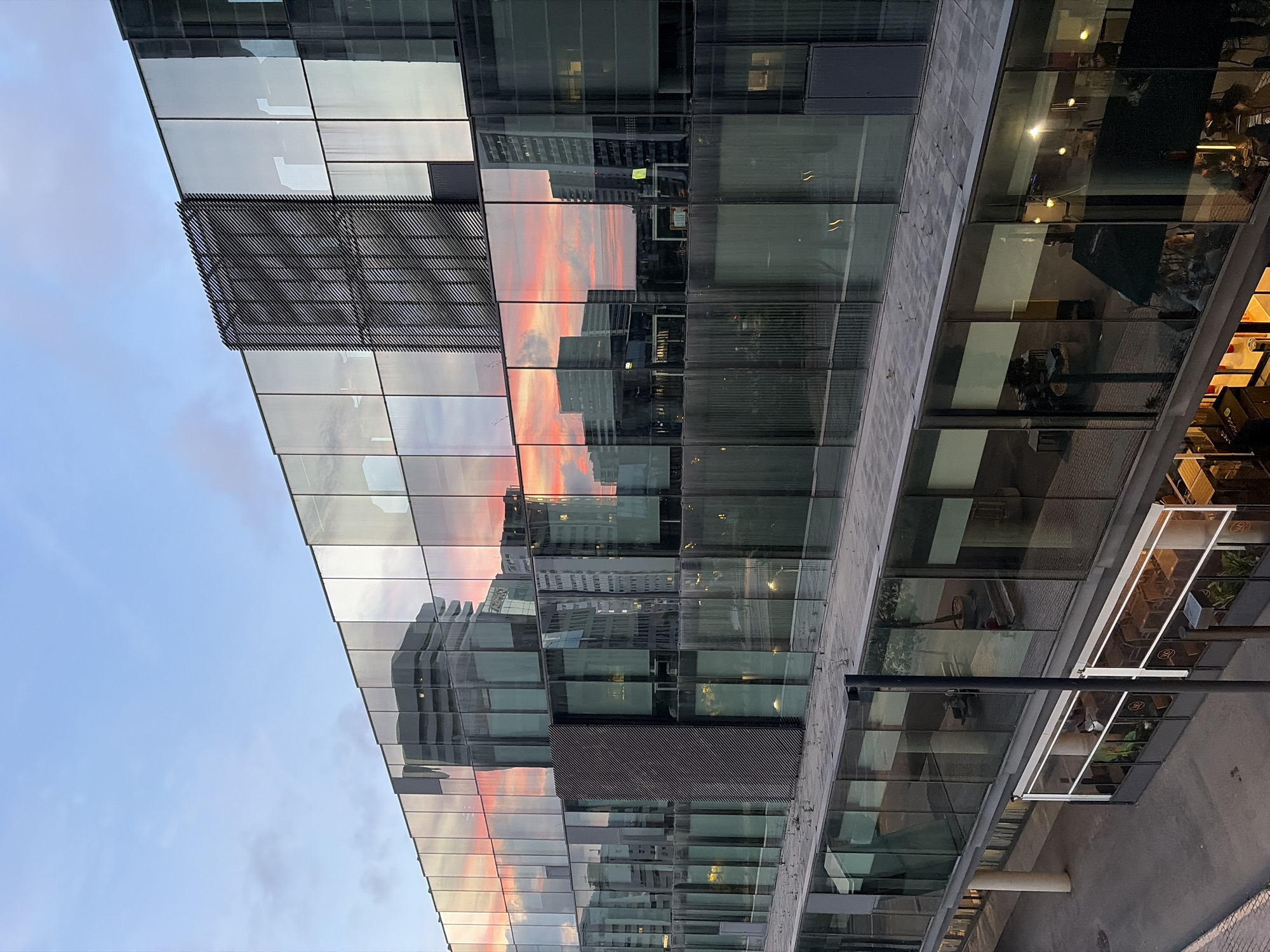 The image displays a modern, multi-story building featuring an extensive glass facade, set in an urban environment in L'hospitalet De Llobregat, Spain. The building's reflective surface is the dominant subject, capturing the dynamic colors of a sunset and reflections of other surrounding structures.
The upper-left portion of the image shows a clear, light blue sky. However, the glass panels of the building prominently reflect a vibrant sunset, characterized by striking hues of orange, pink, and red, stretching across the middle section of the facade. These reflections create a layered visual effect, with silhouettes of distant, taller buildings also visible within the glass.
Architectural details include dark, textured panels with diagonal patterns, appearing as sun shades or decorative elements on parts of the building's exterior. The ground level of the building features large glass windows that reveal an interior space, likely a commercial establishment such as a restaurant or cafe. Warm artificial lighting is visible inside, and faint shapes of people and furniture can be discerned, indicating activity within. A dark vertical pole, possibly part of street furniture or the building's exterior, is present in the lower right foreground.
The overall scene suggests late afternoon or early evening, evidenced by the sunset colors and the illuminated interior spaces. The weather appears clear. No specific event or distinct activity is taking place in the immediate foreground, but the illuminated interior hints at ongoing commercial operations. There is no visible text in the image.