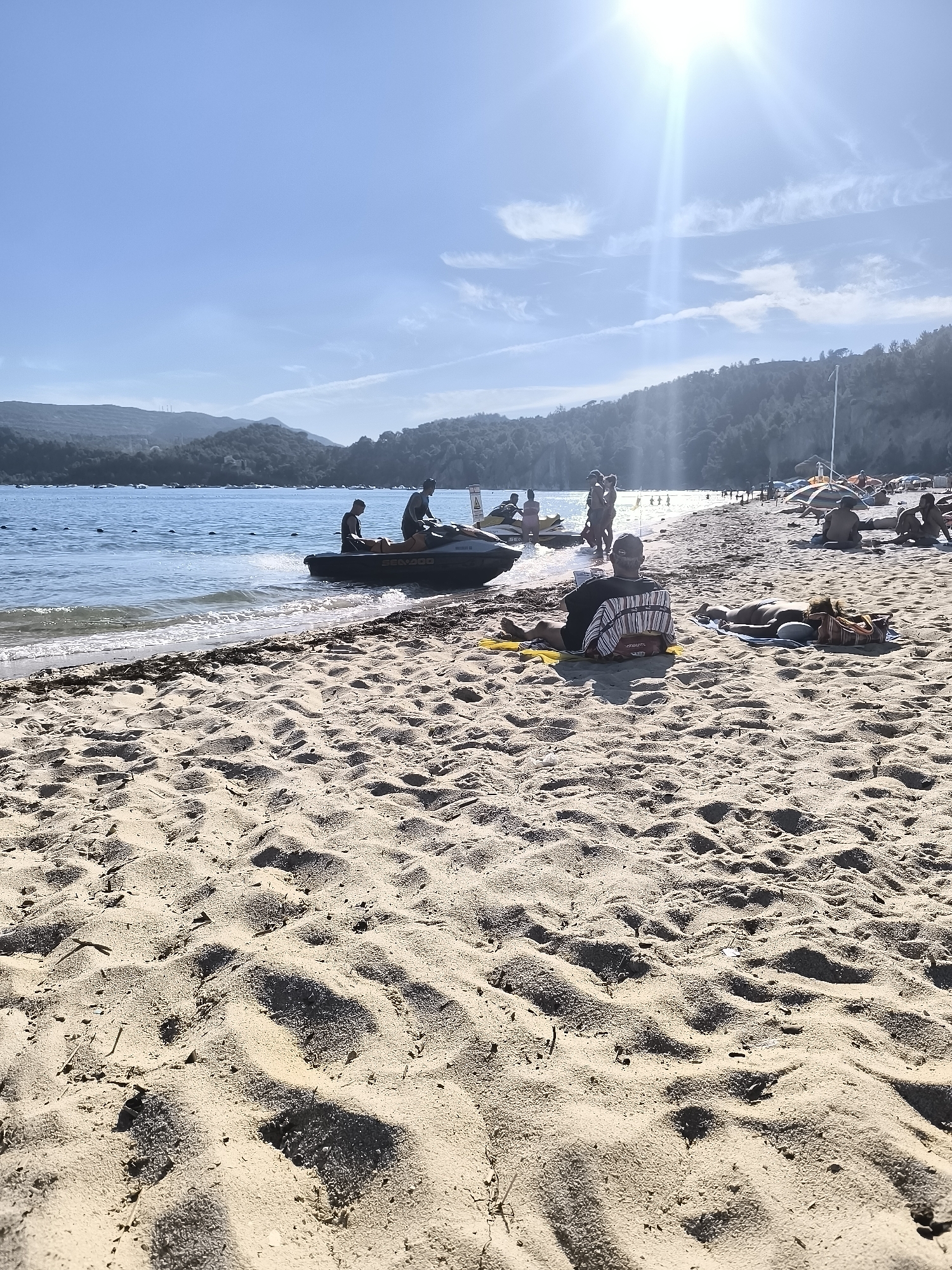 This image depicts a sunny day at a beach, likely in the Setúbal region of Portugal given the hilly, forested coastline and the typical beach activities. In the foreground, the sandy beach is textured with ripples and footprints. Closer to the water, a person sits on a towel, facing away from the camera, wearing a striped shirt and a hat. Beside them, another person lies on their stomach on a towel, sunbathing. In the water, several jet skis are visible. One jet ski is partially beached, with two individuals on it. Further out in the water, other people are engaged in water sports, and more are visible on the beach, with umbrellas and lounging individuals scattered across the sand. The sea is calm, with small waves lapping the shore. The background features a verdant, hilly coastline under a bright blue sky with scattered white clouds and the sun shining brightly.