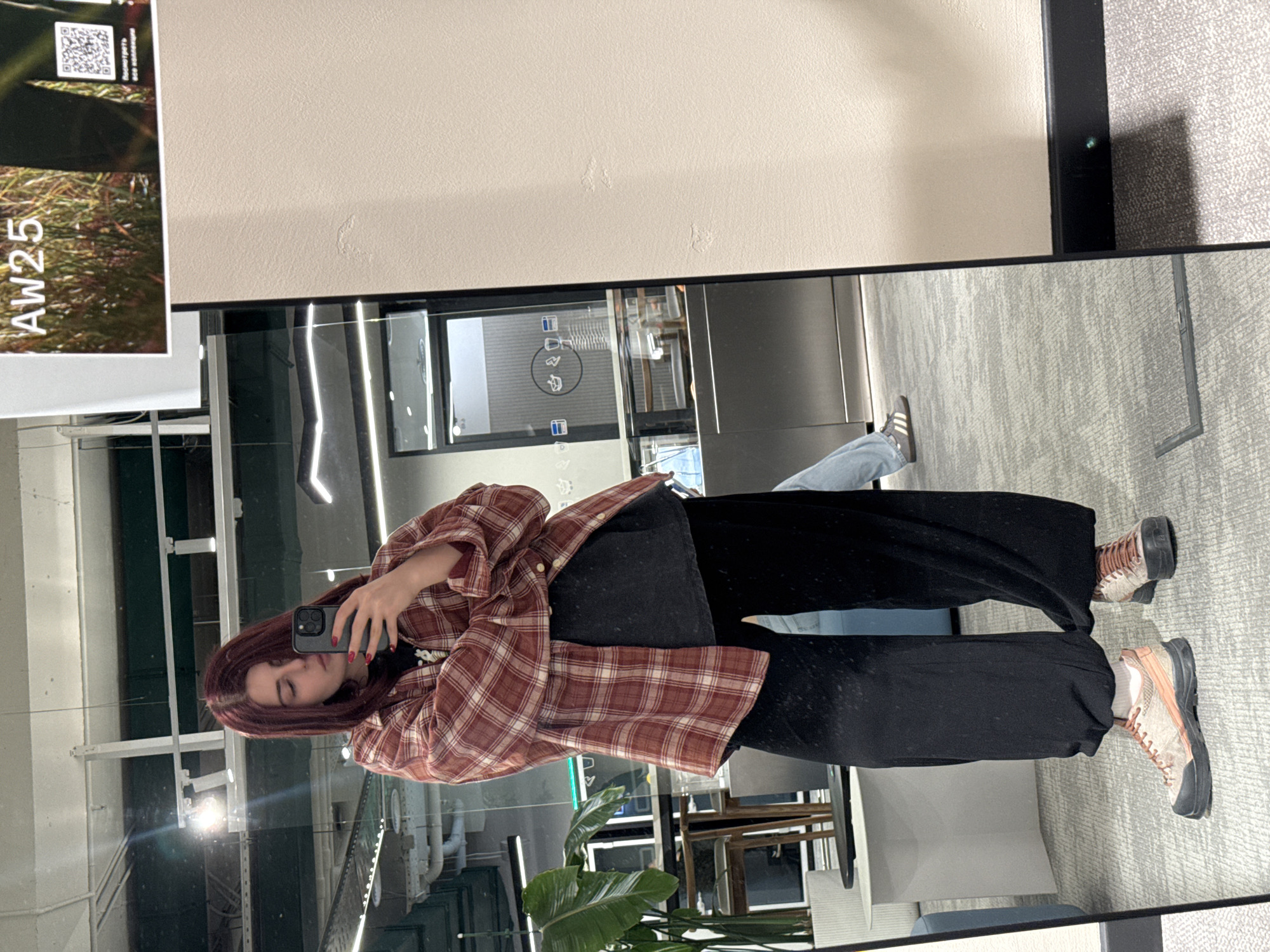A woman takes a selfie in a mirror in what appears to be an office space. The woman has auburn hair and wears a plaid shirt, black pants, and tan hiking boots. A phone is visible in her hand. The reflection in the mirror shows office features, including plants, furniture, and architecture. A document with "Aw25" printed on it is on the left. The scene is possibly in Khimki, Russia, but this cannot be confirmed from the image alone.