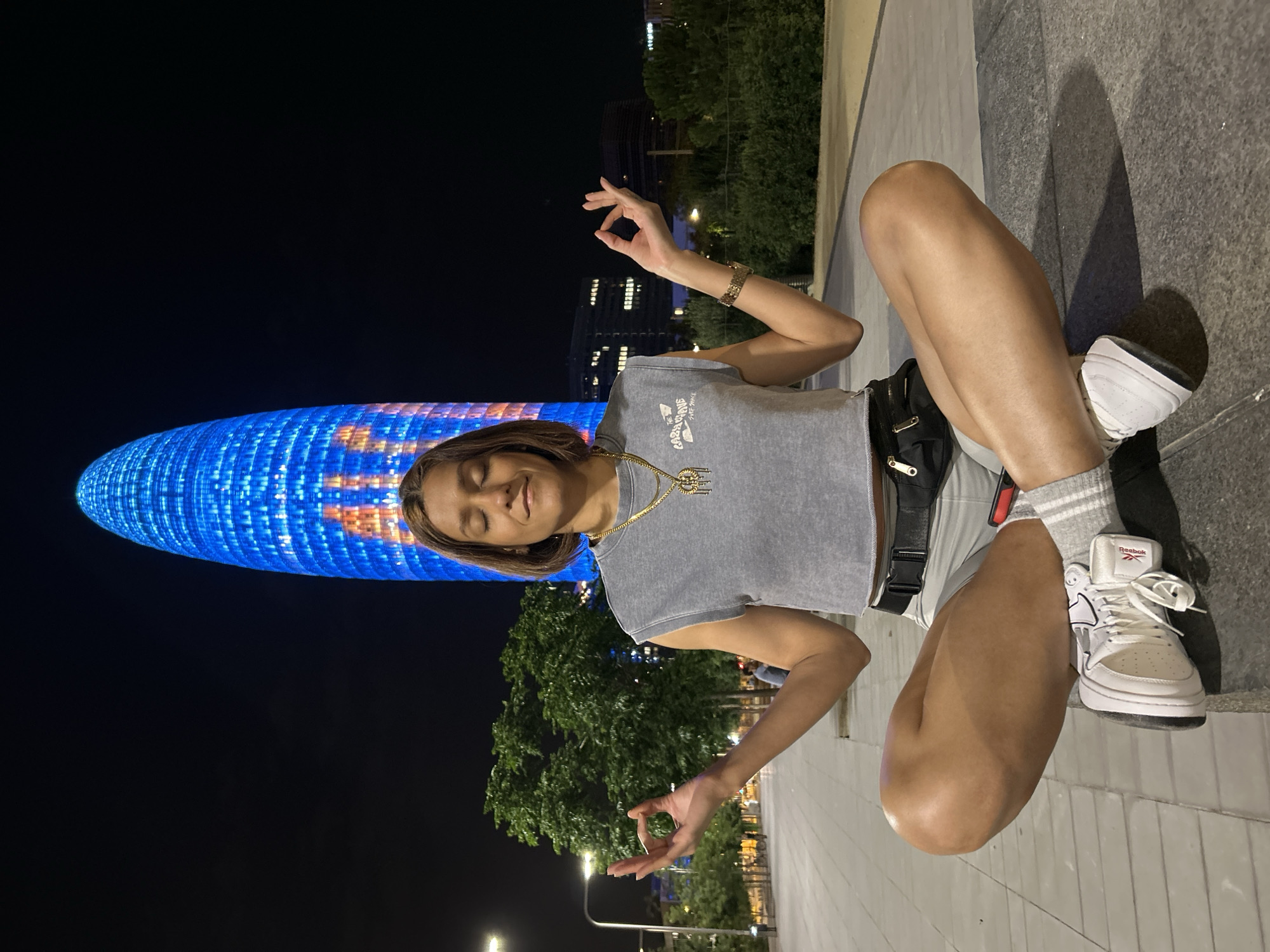 A young woman is meditating in a cross-legged seated position on a stone ledge at night. She is wearing a grey sleeveless crop top, grey shorts, white sneakers with grey and white striped socks, and a black fanny pack around her waist. She has a gold necklace with a pendant. Her eyes are closed and her hands are resting on her knees with her thumbs and index fingers touching, in a meditative gesture. In the background, the Torre Glòries, a 38-story skyscraper in Barcelona, Spain, is illuminated with blue and orange lights. Trees and other buildings are also visible in the background. The ground appears to be paved.