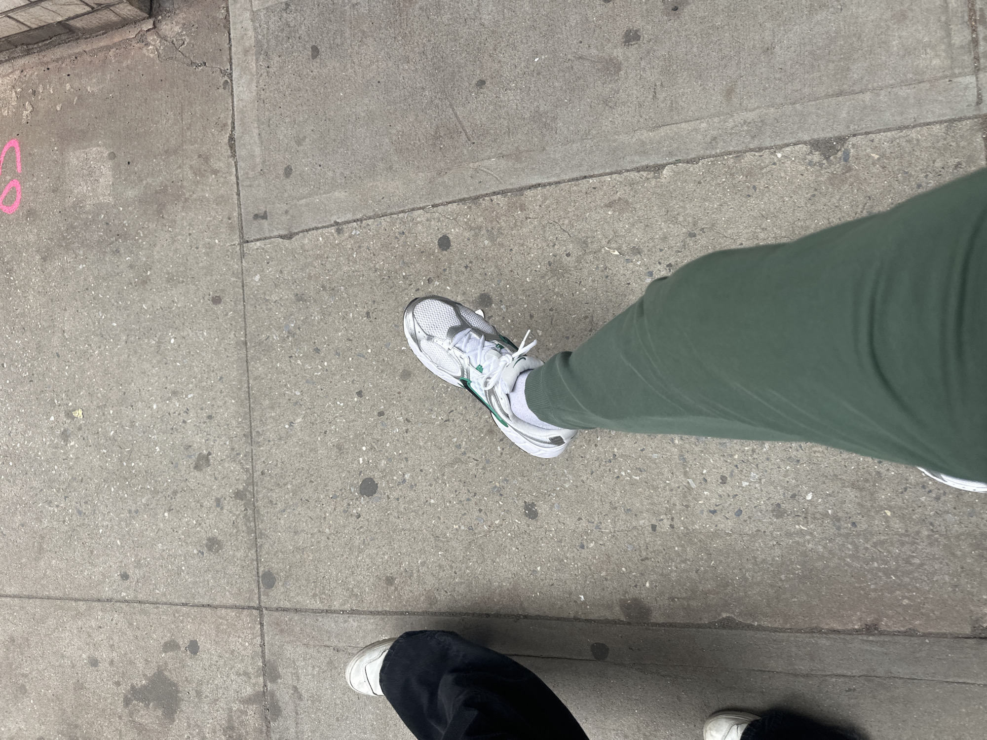 An elevated perspective captures the lower extremities of two individuals on a worn, light gray concrete sidewalk in New York, United States of America. The concrete surface is segmented into rectangular slabs, exhibiting scattered dark stains and minor surface imperfections. A faint, partial pink chalk marking, resembling the upper curve of a digit "2" or an "S", is visible on the upper right slab. The individual on the right wears dark green trousers and white athletic shoes with metallic silver and green accents, along with white socks. Their left foot is positioned centrally, angled forward. The individual on the left wears black trousers and white low-top sneakers, with their lower left leg and foot partially visible along the frame's left edge. The two individuals appear to be standing or walking in close proximity. A brick building foundation or curb is partially visible along the far right edge of the frame.