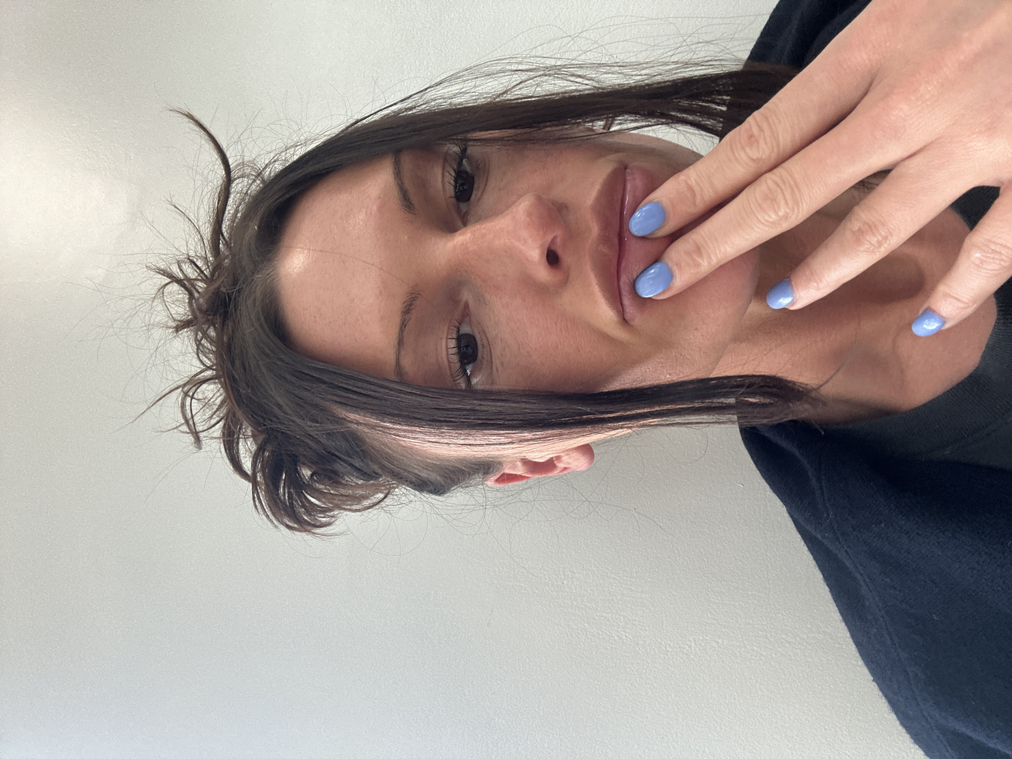 A close-up, portrait-style photograph captures a woman, likely taken as a selfie, with her head tilted to the right. Her gaze is directed towards the camera with a relaxed expression. She has dark hair, styled casually with some flyaways visible around her forehead and sides, and dark eyes with prominent eyelashes. Her right hand is raised, with her fingers resting gently against her lower lip and cheek, partially obscuring her mouth. Her fingernails are painted with a distinct light blue, periwinkle-like polish. She is wearing a dark-colored garment, possibly a black or navy hoodie or top, visible on her right side. The background is a plain, light grey or off-white wall, suggesting an indoor setting. The lighting is soft and even, highlighting her features. No specific time of day or other location cues are visible within the image, though the context provided indicates London, United Kingdom. There is no visible text in the image.