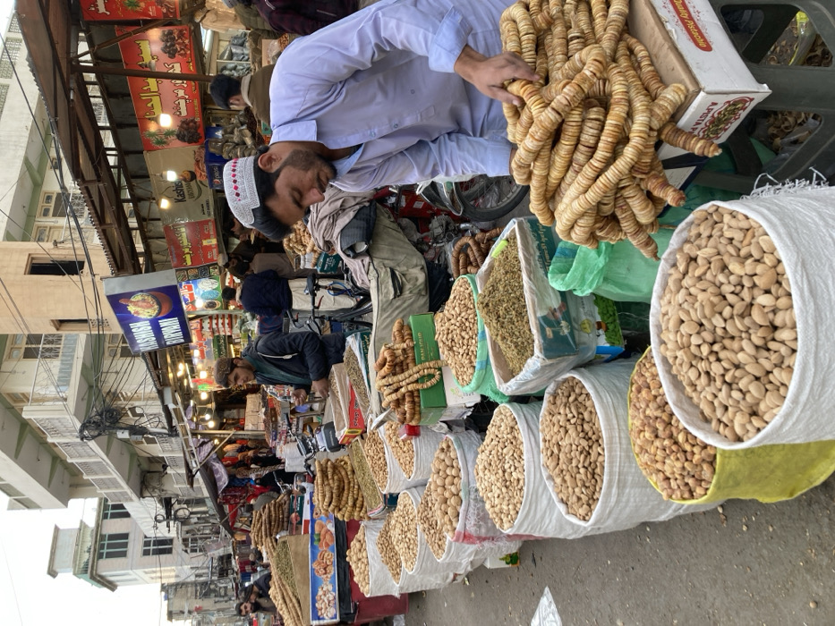 The image depicts an outdoor market scene on a paved surface in Quetta City Tehsil, Pakistan. In the foreground, a linear arrangement of approximately ten large sacks, primarily white but including yellow and green, are filled with various dried nuts and fruits such as almonds, pistachios, walnuts, and peanuts. Rings of dried figs are also prominently displayed on cardboard boxes. A male individual in the immediate right foreground, wearing a light blue shalwar kameez and a white patterned cap, is observed bending forward, manipulating stacked rings of dried figs. Another male individual in the midground-left, dressed in a dark jacket and dark cap, stands behind a stall displaying other dried goods. The background features a busy street with additional market stalls, pedestrians, and parked motorcycles. Multiple signs are visible on the stalls, including a prominent blue sign for "KASHISH BIRYANI". Multi-story concrete buildings with balconies line the street beyond the market. Overhead utility wires crisscross the scene.