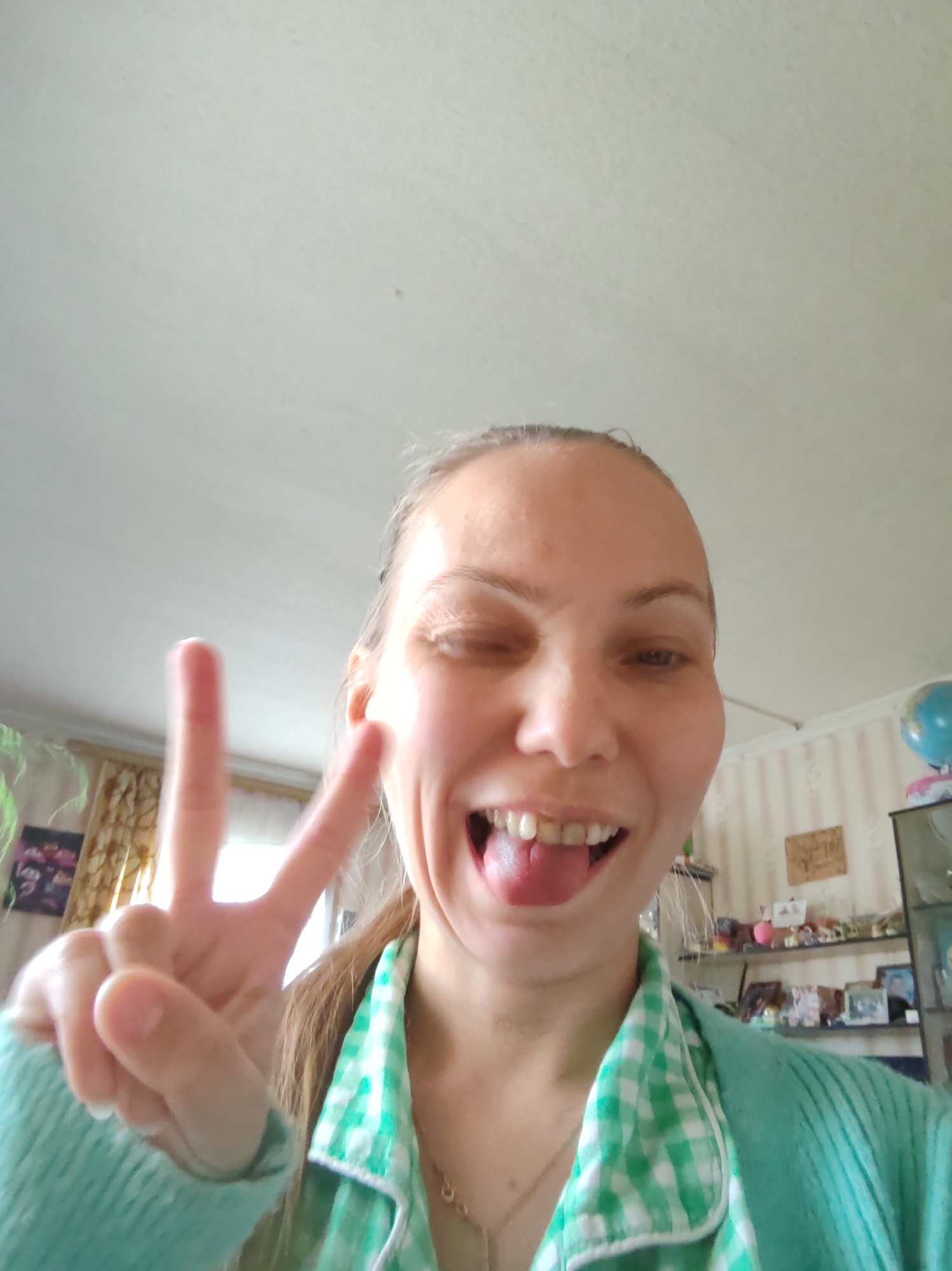 In this close-up, selfie-style shot, a young woman with light brown hair is the sole subject. She is looking directly at the camera, with her right eye closed in a wink and her tongue sticking out playfully. Her mouth is open, revealing her teeth. She is holding up her left hand, forming a peace sign with her index and middle fingers extended.
She is wearing a light blue, textured sweater over a green and white checkered collared shirt. A delicate necklace is visible around her neck. The background suggests an indoor setting, likely a home. A ceiling with visible lighting fixtures is overhead. To the left of the woman, a window with curtains is visible, and a piece of framed artwork hangs on the wall. To the right, a shelf unit is visible, displaying various items including what appears to be a blue globe, framed photographs, and decorative objects. The overall lighting suggests daytime. The woman's expression and gesture convey a lighthearted and cheerful emotion. No text is visible in the image.