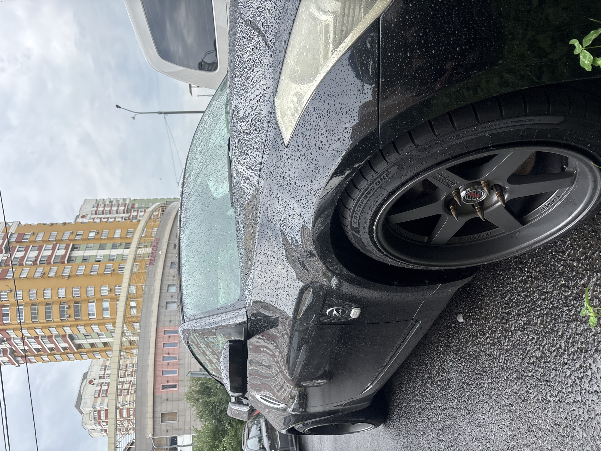 A black Nissan 350Z is parked on a wet asphalt street in Moskovskiy, Russia. The car's surface is covered in raindrops. In the background, a tall, multi-story building with yellow and red accents is visible, along with other buildings and trees. A white van is parked to the right of the Nissan. The sky is overcast.