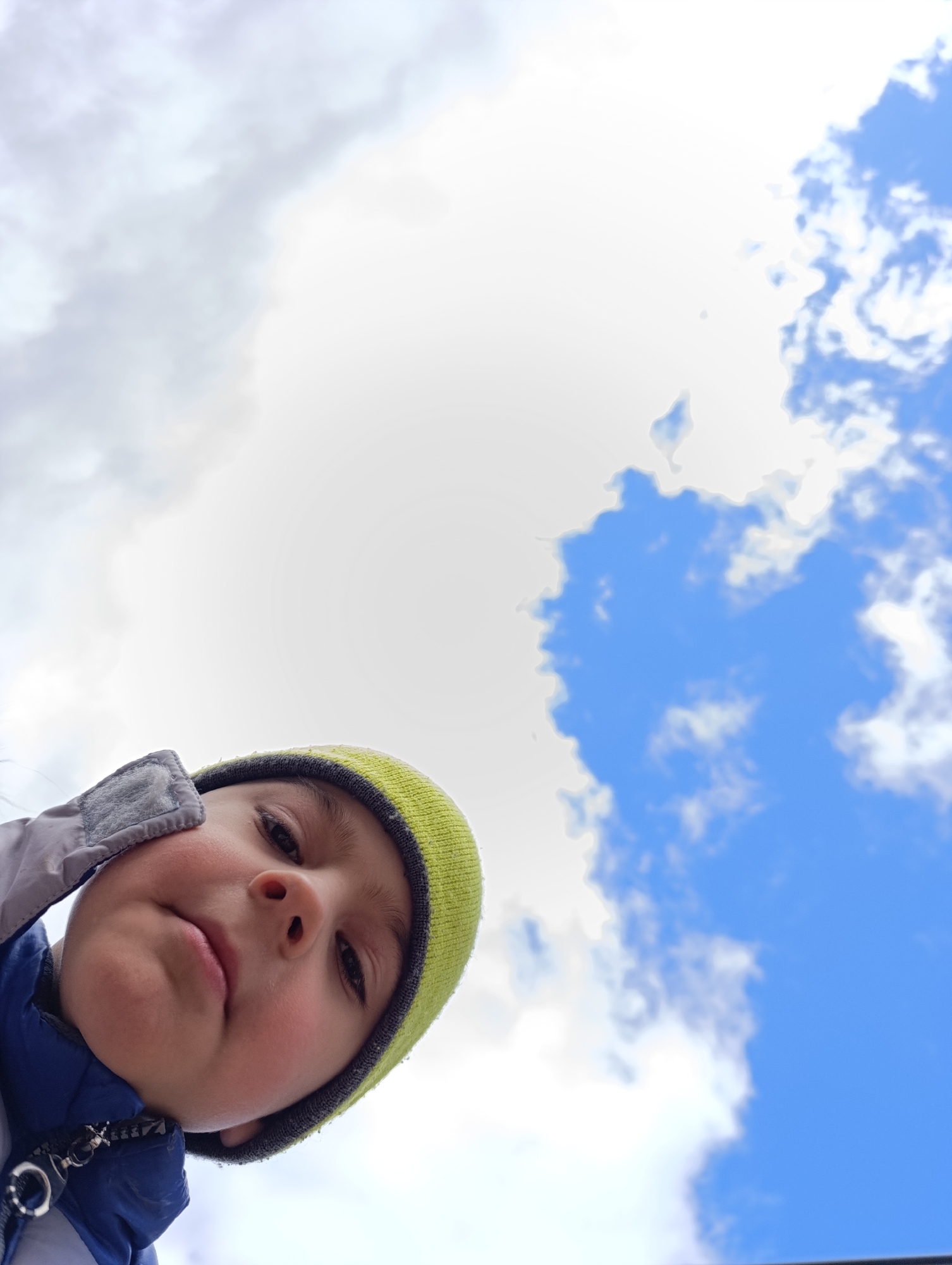 A low-angle close-up captures a young child wearing a light green and dark gray beanie hat, a blue jacket and looking down at the camera. Above the child, the sky is visible with both white and blue, and fluffy clouds. The location, Chaikovsky, Russia, cannot be determined.