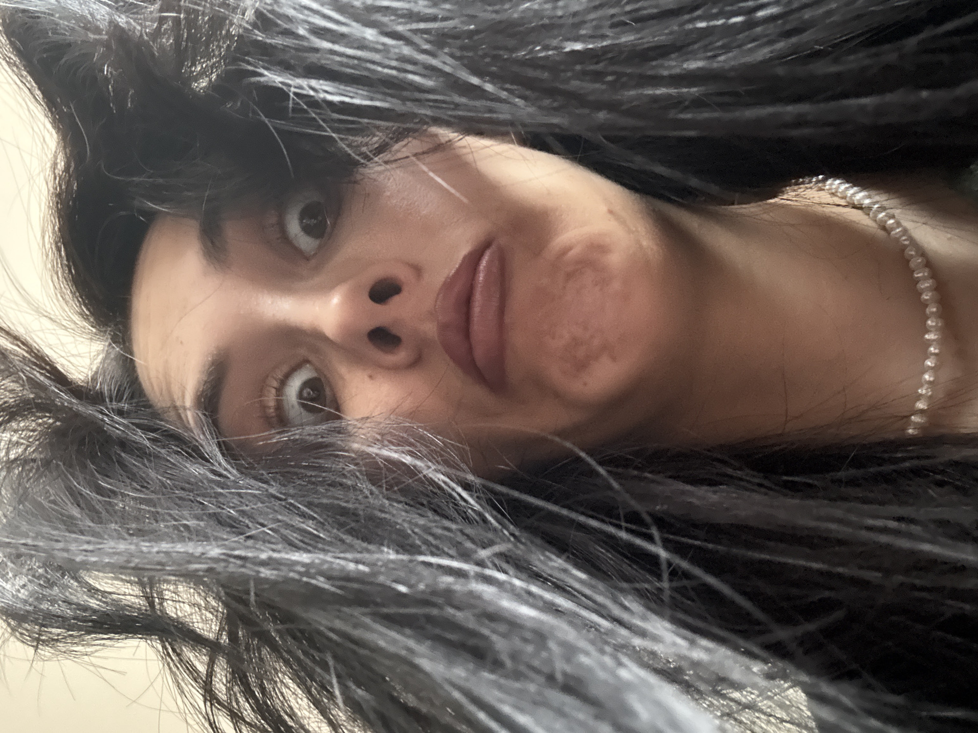 A close-up, rotated portrait features a person with long, dark hair looking directly into the camera. The person's eyes are wide, and their mouth is slightly open, conveying an expression of curiosity, surprise, or playfulness. Dark, seemingly disheveled hair frames the face, with strands falling around and partially across it; some lighter, possibly graying, strands are visible within the dark hair. The skin on the person's right cheek shows visible marks or discolorations. A simple white pearl necklace is visible around the neck, partially covered by hair. The background is a plain, light color, suggesting an indoor setting. The image captures an intimate and candid moment. No text is visible in the media file. The location context for this image is Votkinsk, Russia.