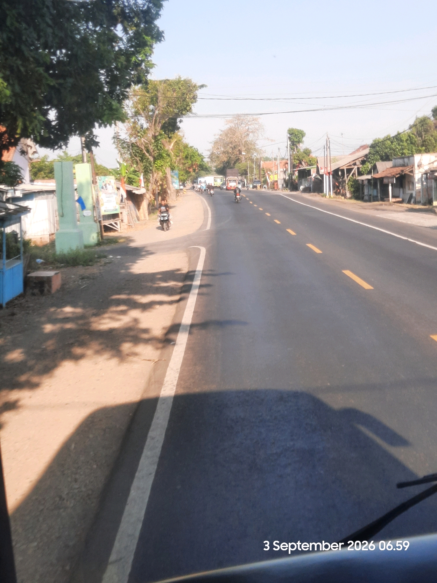 The image captures a road scene in Bulang, Indonesia, on September 3, 2026, at 06:59. A paved road with yellow dashed lines runs through the frame, bordered by a dirt shoulder on the left and buildings on the right. Trees, some with dense foliage, line the left side of the road, casting shadows on the shoulder. Several structures, including a blue stall and a light green pillar, are visible on the left. On the right, a row of buildings with various roof types, some appearing to be shops or houses, are situated along the roadside. A few motorcycles are present on the road, with one rider clearly visible in the foreground on the left. The sky is clear and bright, indicating daylight. A portion of a vehicle, possibly a car or bus, with a windshield wiper, is visible in the lower foreground.