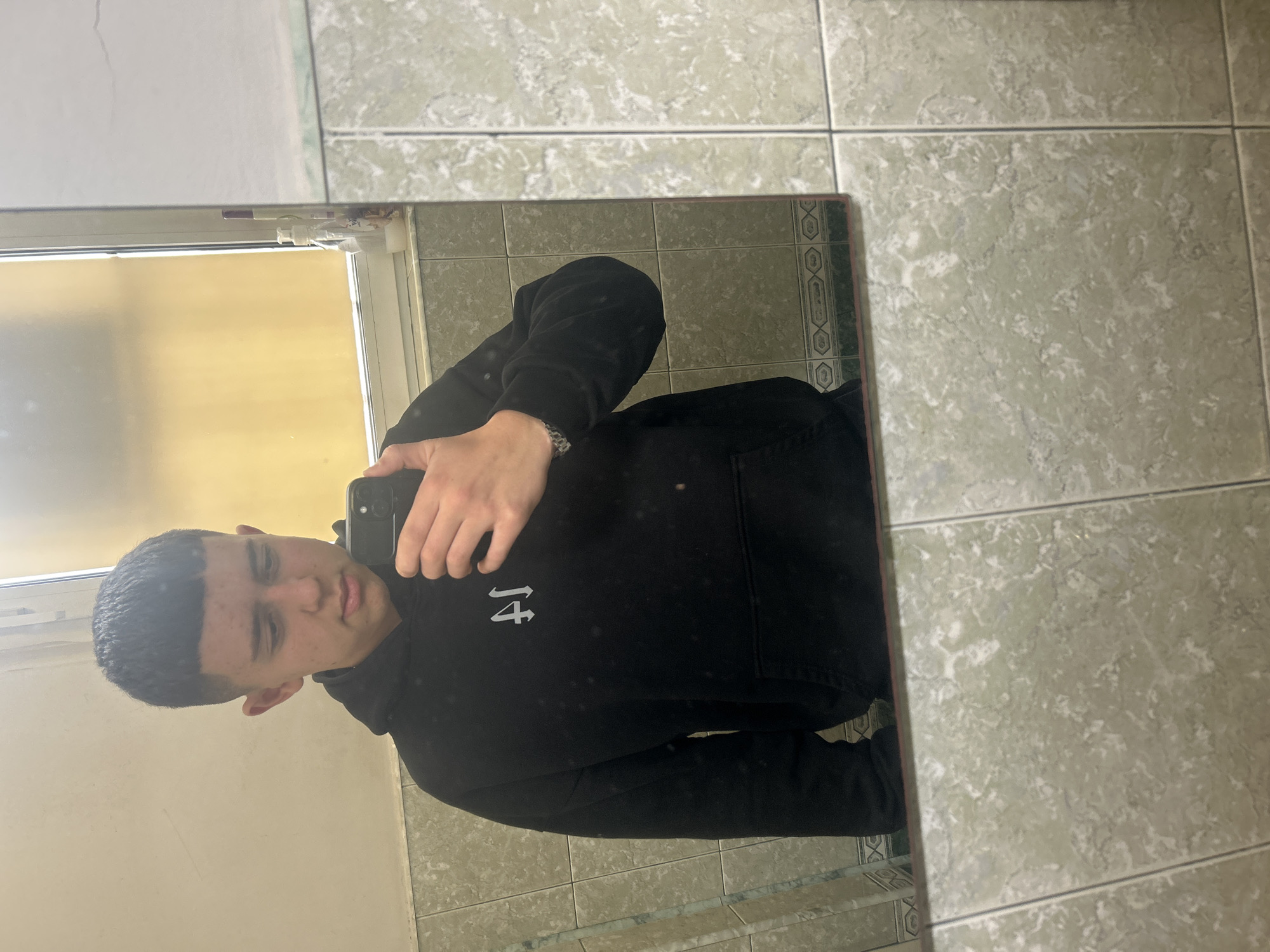 A young man is taking a selfie in a mirror. He is wearing a black hoodie with a white symbol on the left side. The background consists of light green tiles, a window, and a white frame around the window. The photograph was taken in Zlatograd, Bulgaria.
