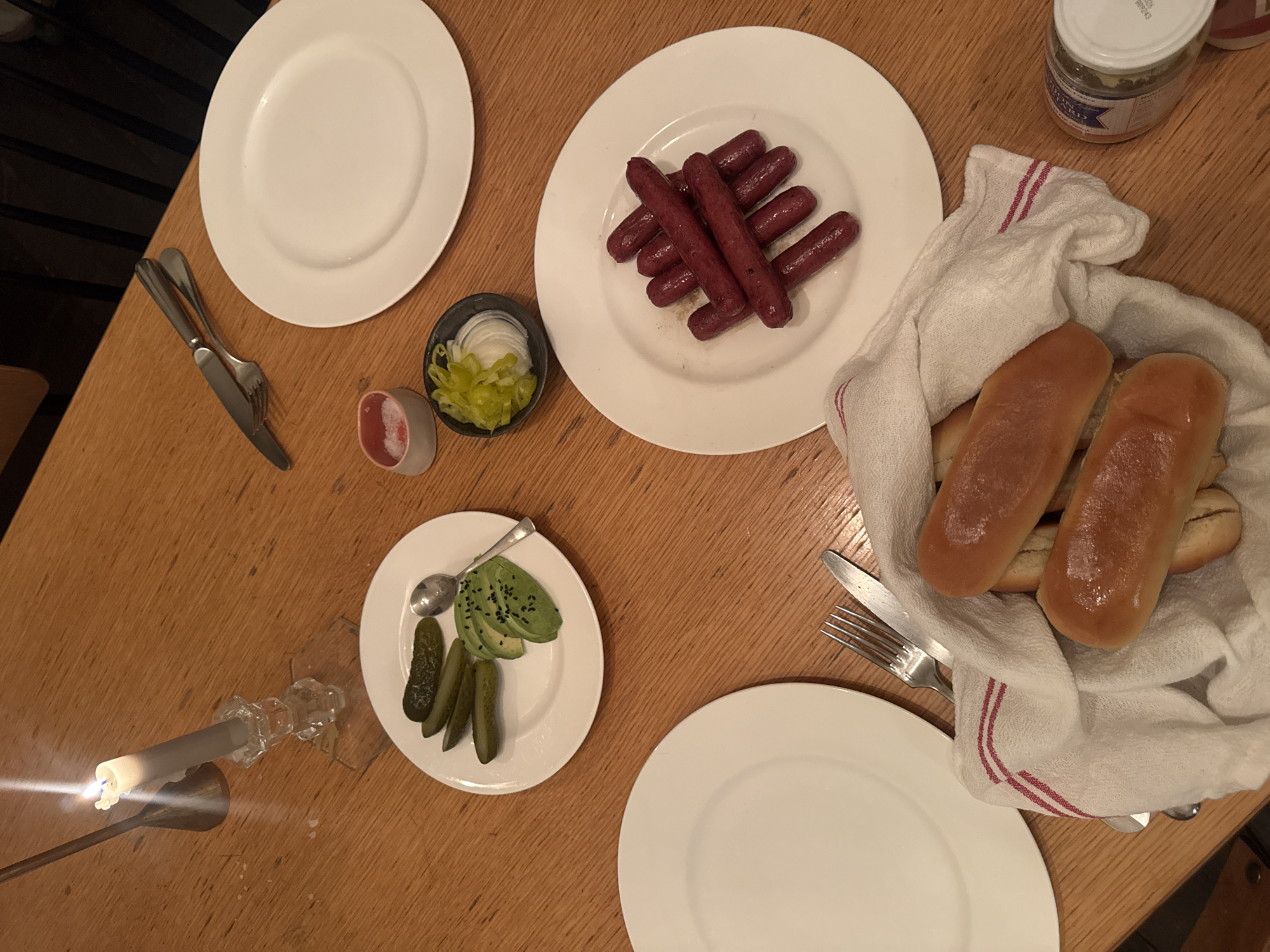 A table in New York City, United States, is set with plates of food. There are hot dogs on one plate, hot dog buns in a cloth-lined basket, and pickles and sliced avocado on another. Condiments and sliced onions are also visible. A lit candle provides illumination.