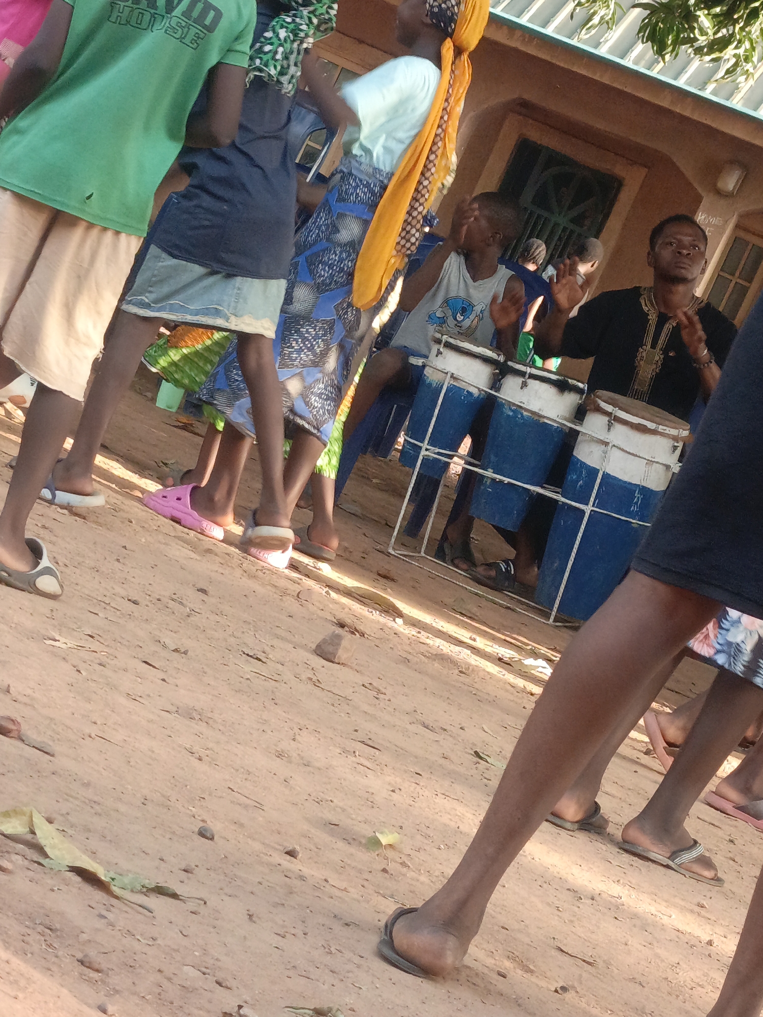 The image shows a gathering of people in Akwanga, Nigeria. A man in a black shirt with gold embroidery is playing a set of three blue and white drums. A younger person wearing a tank top is seated near him. Several other individuals stand around them, dressed in various clothing including skirts, shorts, and a green shirt with the word "HOUSE" visible. Some people are wearing sandals. The ground is reddish-brown dirt. A building is visible in the background.