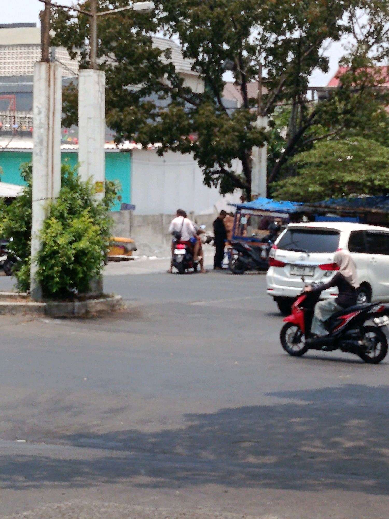 In an outdoor scene in the city of Unknown, Indonesia, a street intersection is depicted with several elements of urban life. A white SUV is parked on the right side of the image, and a red and black motorcycle is in motion to the left of the SUV. A person wearing a hijab is riding the red and black motorcycle. To the left of the SUV, a person on a black motorcycle is partially visible, and further left, another motorcycle is parked. In the background, there are trees, a building with a white facade and a tiled roof, and a blue awning over what appears to be a food stall. The ground is paved, suggesting a road or parking area.