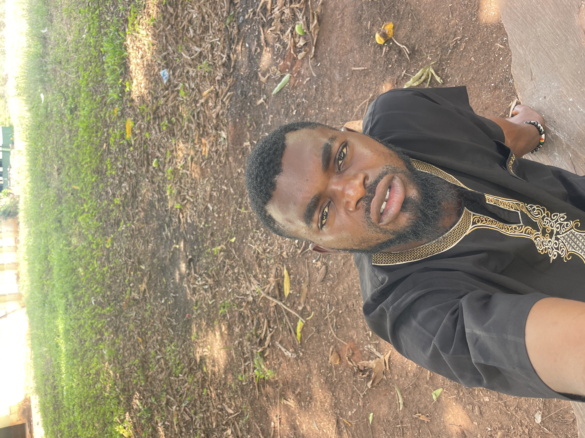 A man with dark skin, a beard, and short hair is posing for a selfie outside. He is wearing a black shirt with gold embroidery around the collar. He is wearing a beaded bracelet on his right wrist. The ground is a mix of grass and dirt with leaves and twigs scattered around. The location is Ogbe, Nigeria.