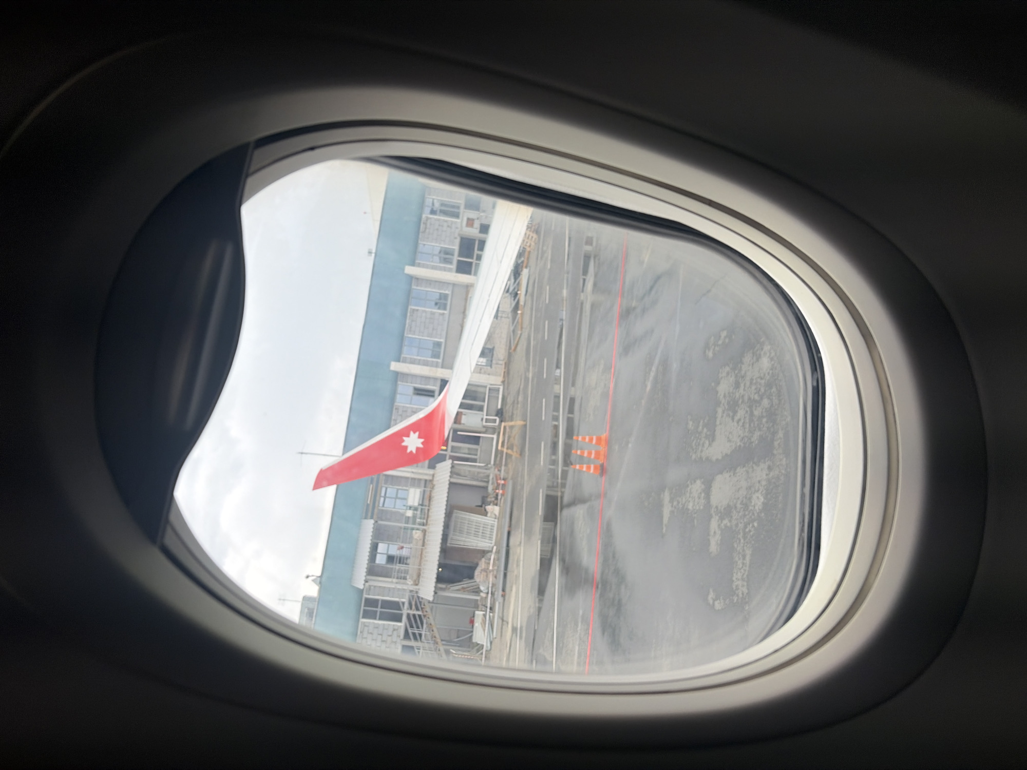 The image offers a view from inside an aircraft cabin, looking out through a porthole window. The light gray window frame defines the foreground, framing the scene outside. A portion of a white airplane wing is visible, extending into the frame from the left. Attached near the wing's edge is a distinct red flag featuring a white seven-pointed star in its center.
Beyond the wing, the airport tarmac is visible, appearing to be a dark, possibly wet, surface with painted white and red lines marking boundaries or pathways. Three bright orange traffic cones are strategically placed on the ground. In the background, several multi-story buildings with numerous windows line the airport, some appearing to be under construction or renovation, indicated by scaffolding-like structures. The sky overhead is bright but overcast, suggesting a cloudy daytime.
The scene depicts an aircraft parked on an airport apron in Новое Мартьяново, Russia. No people are visible, and there is no discernible text on any objects.