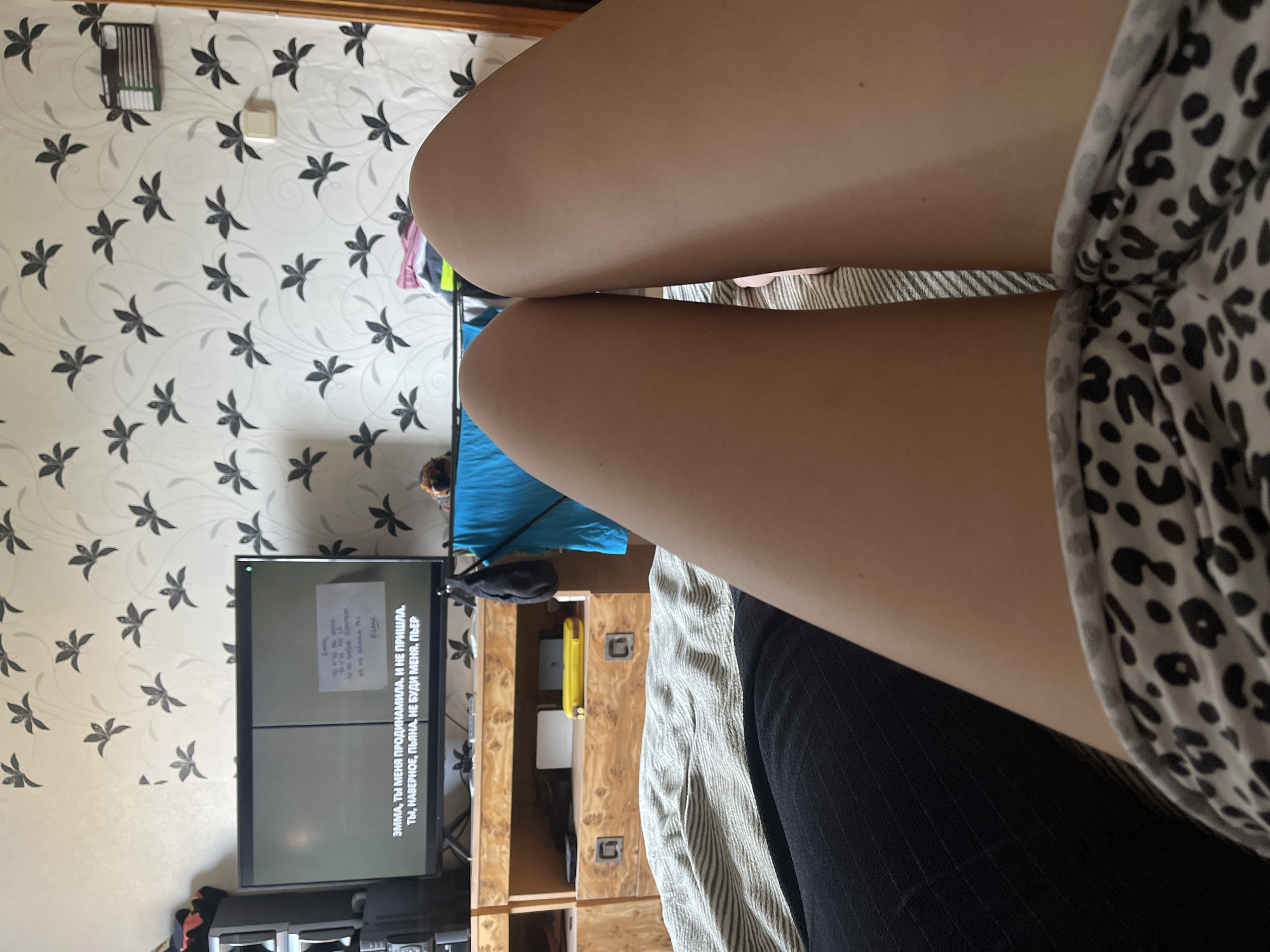 The image shows a close-up view of a person's legs, with their thighs prominently featured. The person is wearing a white top with a black leopard print pattern. The background consists of a wall with a black floral wallpaper pattern, a television displaying text in Russian, and a wooden cabinet. The TV screen shows the text: "ЭММА, ТЫ МЕНЯ ПРОДИНАМИЛА. И НЕ ПРИШЛА. ТЫ, НАВЕРНОЕ, ПЬЯНА. НЕ БУДИ МЕНЯ. ПЬЕР". The setting appears to be indoors, likely a living room, based on the presence of the TV and cabinet. The lighting suggests it is daytime. The overall impression is a casual, indoor scene focused on the person's legs and the text on the screen. There are no discernible external environmental cues.