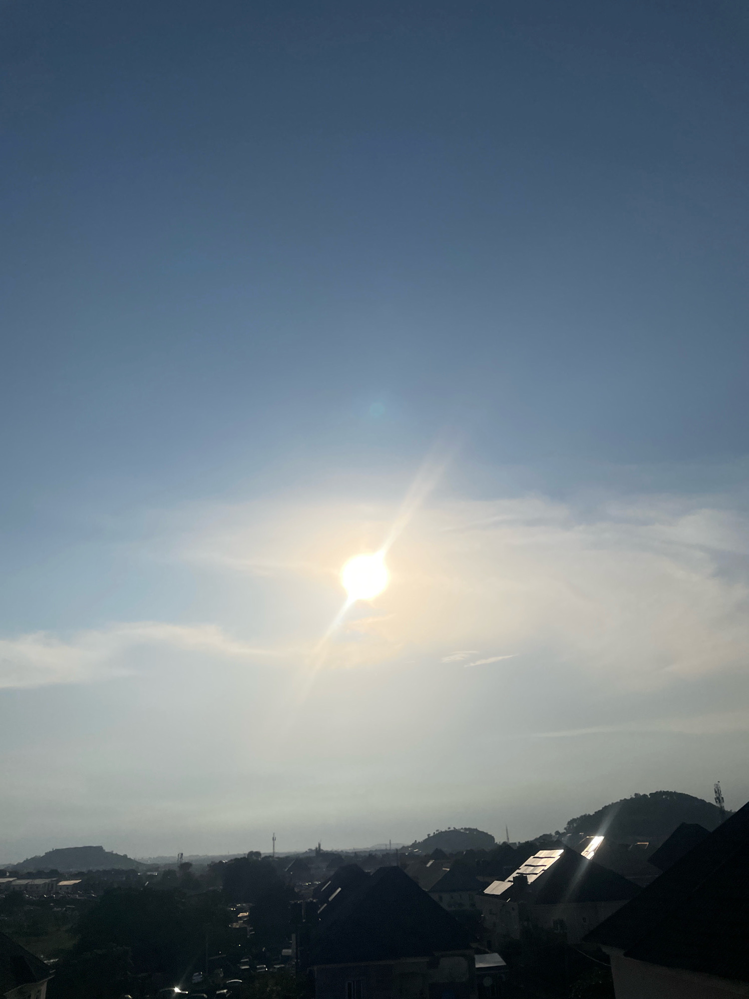 The image captures a panoramic view of a cityscape, likely within Abuja, Nigeria, under a bright, hazy sky. The sun is prominently positioned in the upper center, radiating a strong glare with visible light flares. Thin, wispy clouds are scattered across the sky, partially obscuring the sun.
In the midground and foreground, the scene is dominated by a dense collection of buildings with dark-colored, angled roofs, suggesting a residential or commercial area. Some of these roofs exhibit strong reflections of sunlight, indicating the angle of the sun and the materials of the roofing. Trees are interspersed among the structures, with some areas appearing more densely wooded.
In the distance, rolling hills or mountains form a backdrop to the city. The overall atmosphere is slightly diffused, possibly due to the haze or the time of day. The image appears to be taken from an elevated perspective, looking down onto the city. No individuals are visible in the frame. The scene is still and quiet, depicting a typical urban landscape under a sunny, possibly humid, climate.