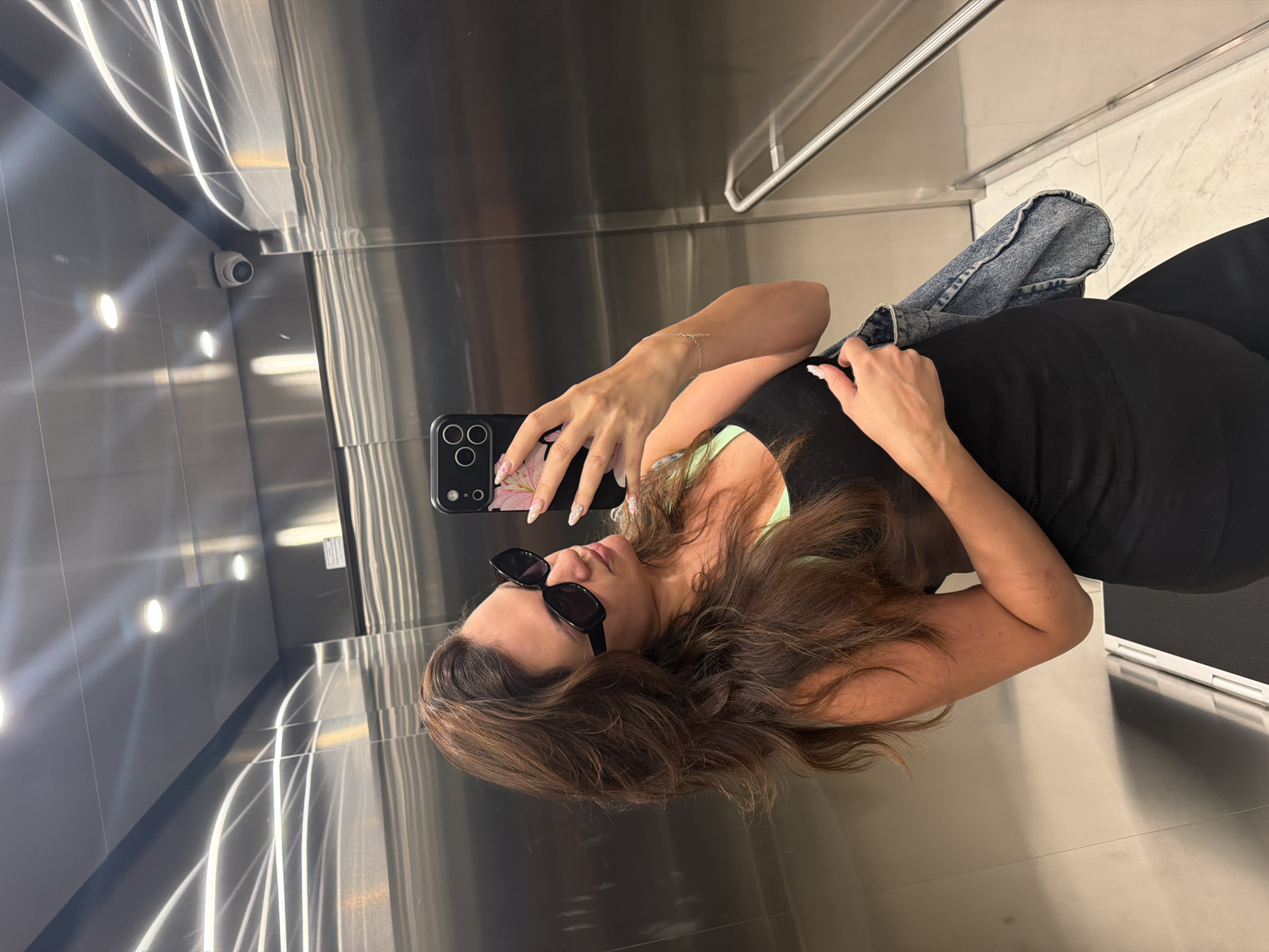 A woman is taking a selfie in an elevator. She is wearing sunglasses, a black tank top with a lime green bra underneath, and black leggings. She is holding a denim bag in her left hand and her phone in her right hand. The elevator has stainless steel walls and a polished floor. There is a security camera visible on the ceiling. The woman has long, wavy brown hair. She is wearing a bracelet on her left wrist and has manicured nails with a floral design. The scene appears to be in an indoor setting, possibly a hotel or residential building. There is no indication of the specific location within Belgrade, Serbia.