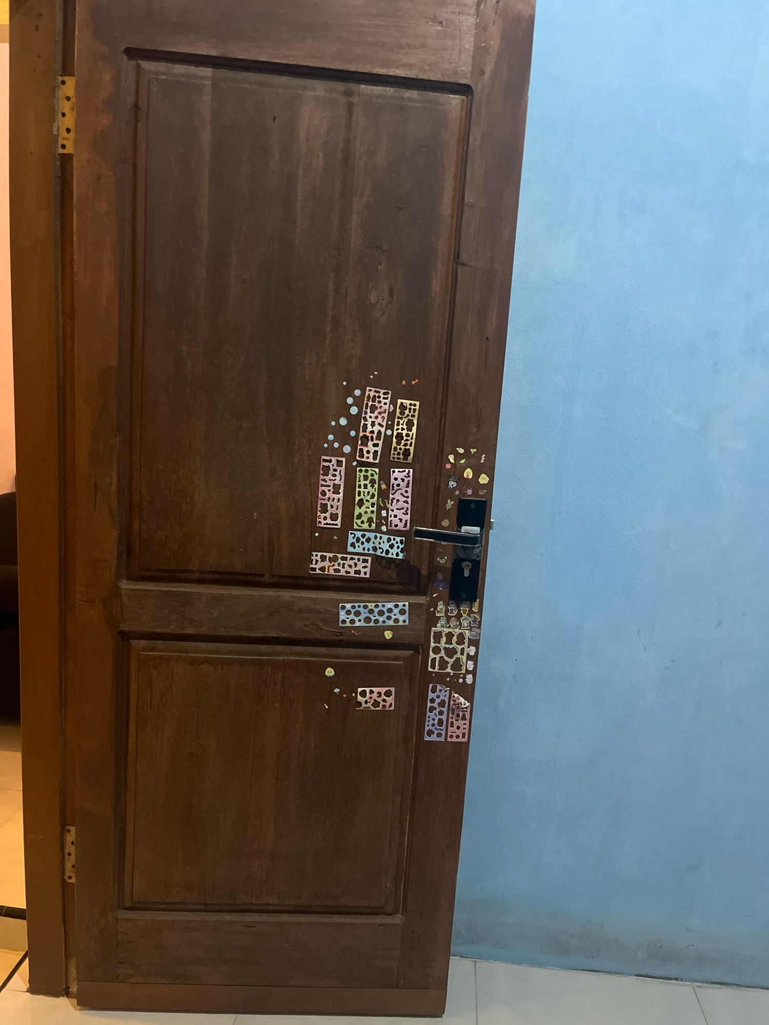 A wooden door in a room in Kebumen, Indonesia, has several colorful stickers applied to it, some of which are patterned and perforated. The door is dark brown and appears to be aged. A silver door handle with a black grip is attached to the right side of the door. The door is slightly ajar, revealing a sliver of a cream-colored wall and a dark interior to the left. The background to the right of the door is a light blue wall. The floor is tiled with white and gray squares. The image is taken from a medium distance.