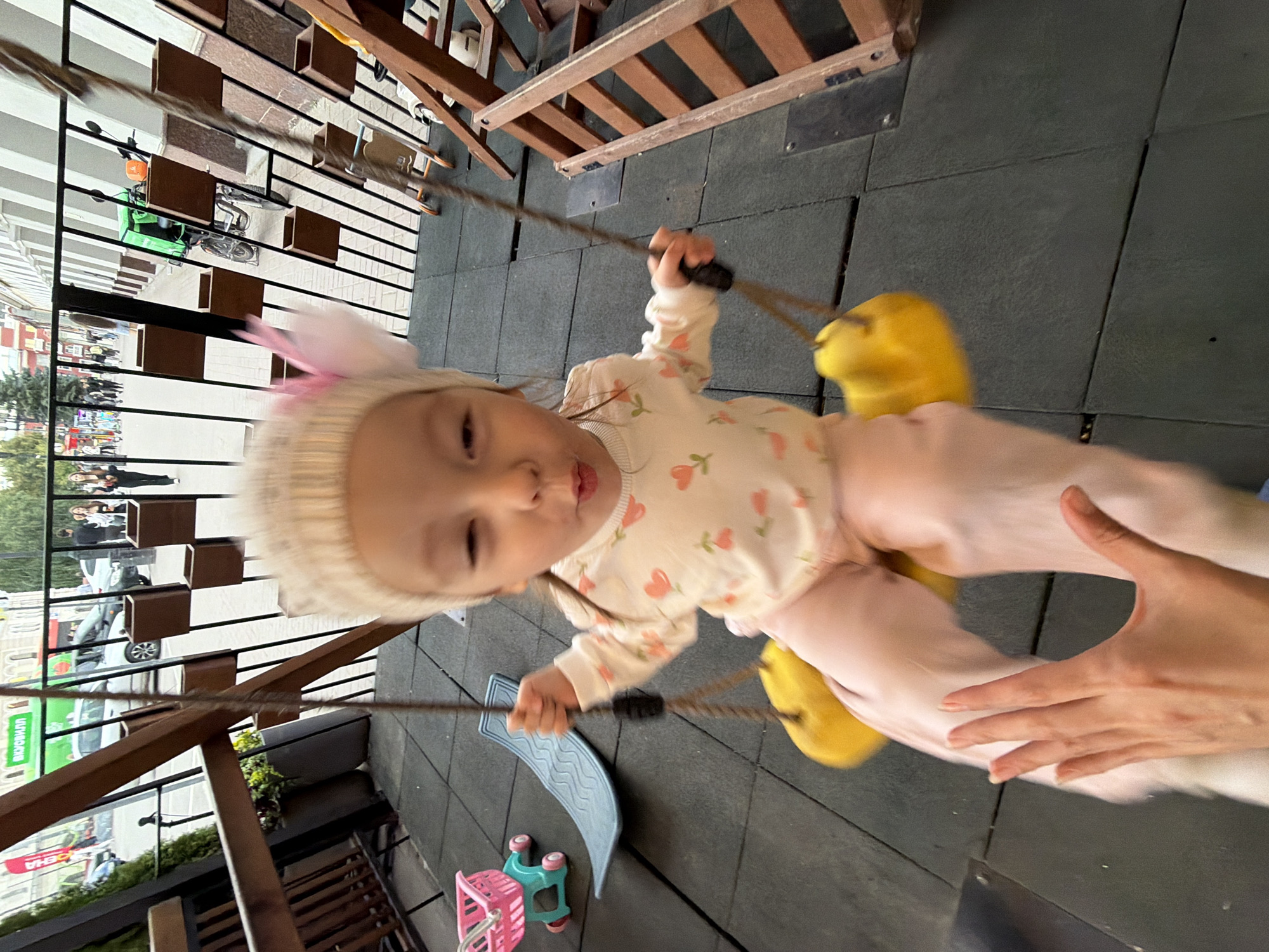 A young child, wearing a pink and white patterned sweater and a cream-colored knitted hat with a pink bow, is on a yellow swing. The child's mouth is puckered. The swing is suspended by ropes. In the foreground, a person's hand, with fingernails painted red, is visible reaching out towards the child. Behind the child, a blue plastic slide and a pink toy shopping cart are on the ground. The ground is made of interlocking black rubber tiles. In the background, a metal fence with wooden planters is visible, and beyond that, a street with buildings, cars, and pedestrians can be seen. This scene appears to be an outdoor playground area, possibly part of a cafe or public space in Ufa, Russia, given the presence of Cyrillic text on a green banner in the background.