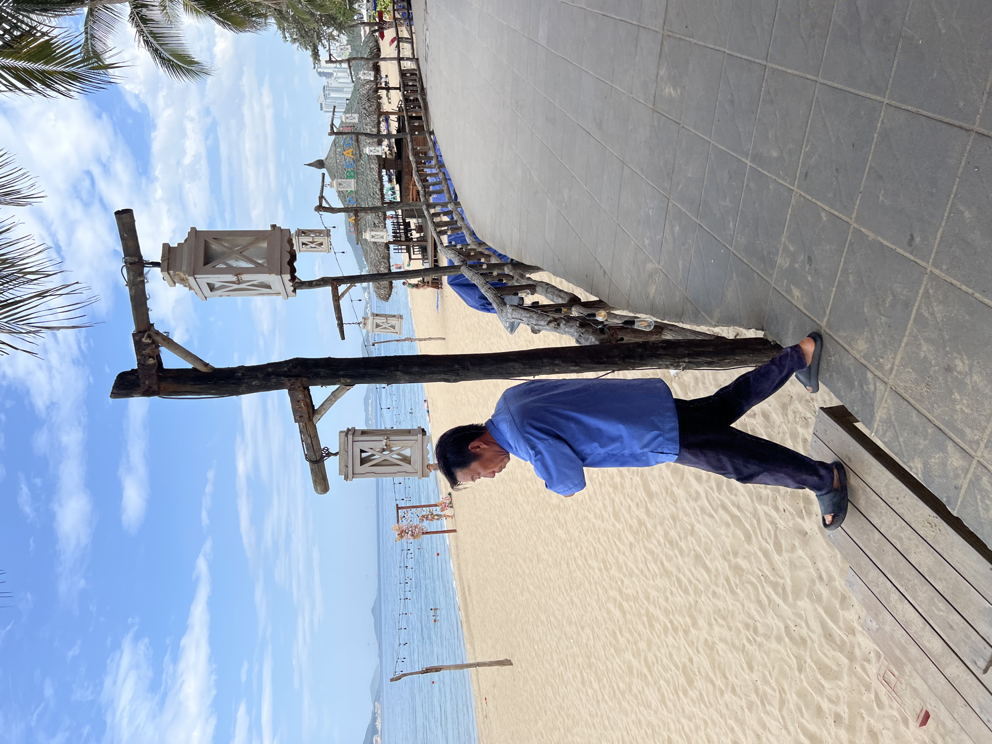 An Asian man in a blue button-down shirt and dark pants walks on a wooden walkway next to a sandy beach in Nha Trang, Vietnam. The man is walking towards the left of the frame and is looking down. Behind him, the beach stretches out to the ocean, with several palm trees visible. In the background, there are thatched-roof structures and buildings, suggesting a resort or dining area. The sky is blue with scattered clouds. The image appears to be taken on a sunny day.