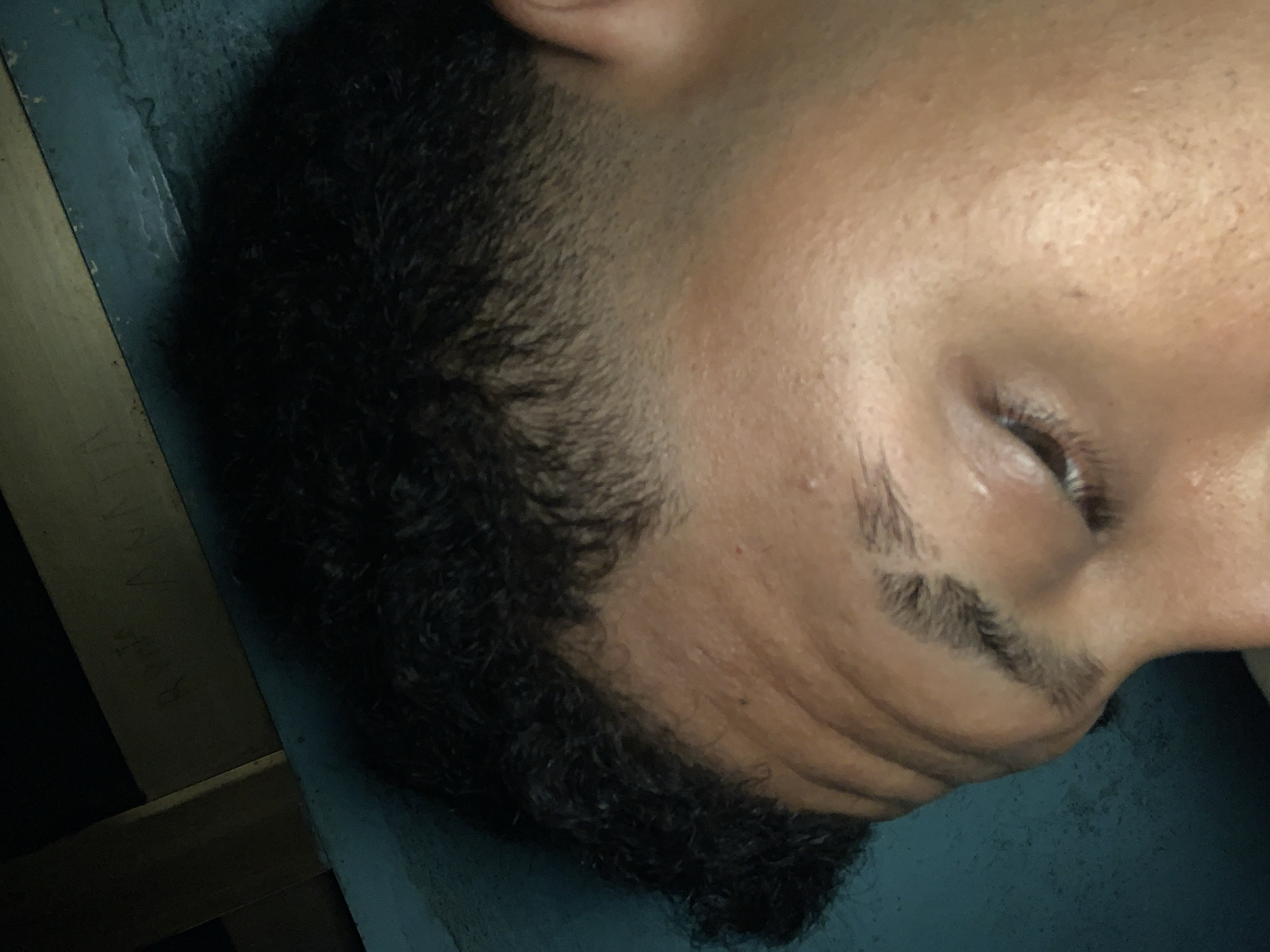 The image captures a close-up, angled side profile of a person's head, appearing to be resting or sleeping. The subject has dark, curly hair styled with a fade haircut. Their eye is closed or cast downwards, revealing dark eyelashes and a thick eyebrow. Visible wrinkles are present on the forehead and the side of the face. The skin is of medium tone, showing visible pores and a small blemish on the temple area.
The person is positioned against a light blue or teal surface, possibly a pillow or fabric, which is seen in the lower part of the frame. In the background, to the upper left, a wooden beam is visible. On this beam, faint, handwritten text can be partially discerned, including what appears to be "RIVAN" among other less legible marks. The lighting is dim, casting soft shadows that highlight facial contours and hair texture. The scene suggests an indoor setting, and the person appears to be in a state of rest or sleep. No specific location cues are visible within the image to tie it to Santa Ana, Venezuela, other than the provided context.