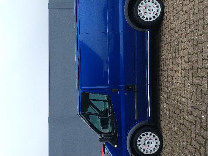 A royal blue Ford Transit panel van is depicted in profile, parked on a paved surface. The vehicle, observed from its driver's side, features dark grey plastic wheel arch extensions and white multi-spoke alloy wheels with red "SPEEDLINE CORSE" lettering visible on the front wheel. A chrome-colored trim strip runs along the lower side of the bodywork. The "TRANSIT" emblem is visible on the driver's door. The ground is paved with interlocking concrete bricks in a herringbone pattern, displaying earth tones and some moss growth. In the background, a plain dark grey wall, possibly concrete or rendered, extends horizontally. Above the wall, an overcast, light grey sky is visible. No individuals or active subjects are present. The scene is situated in Saarlouis, Germany.