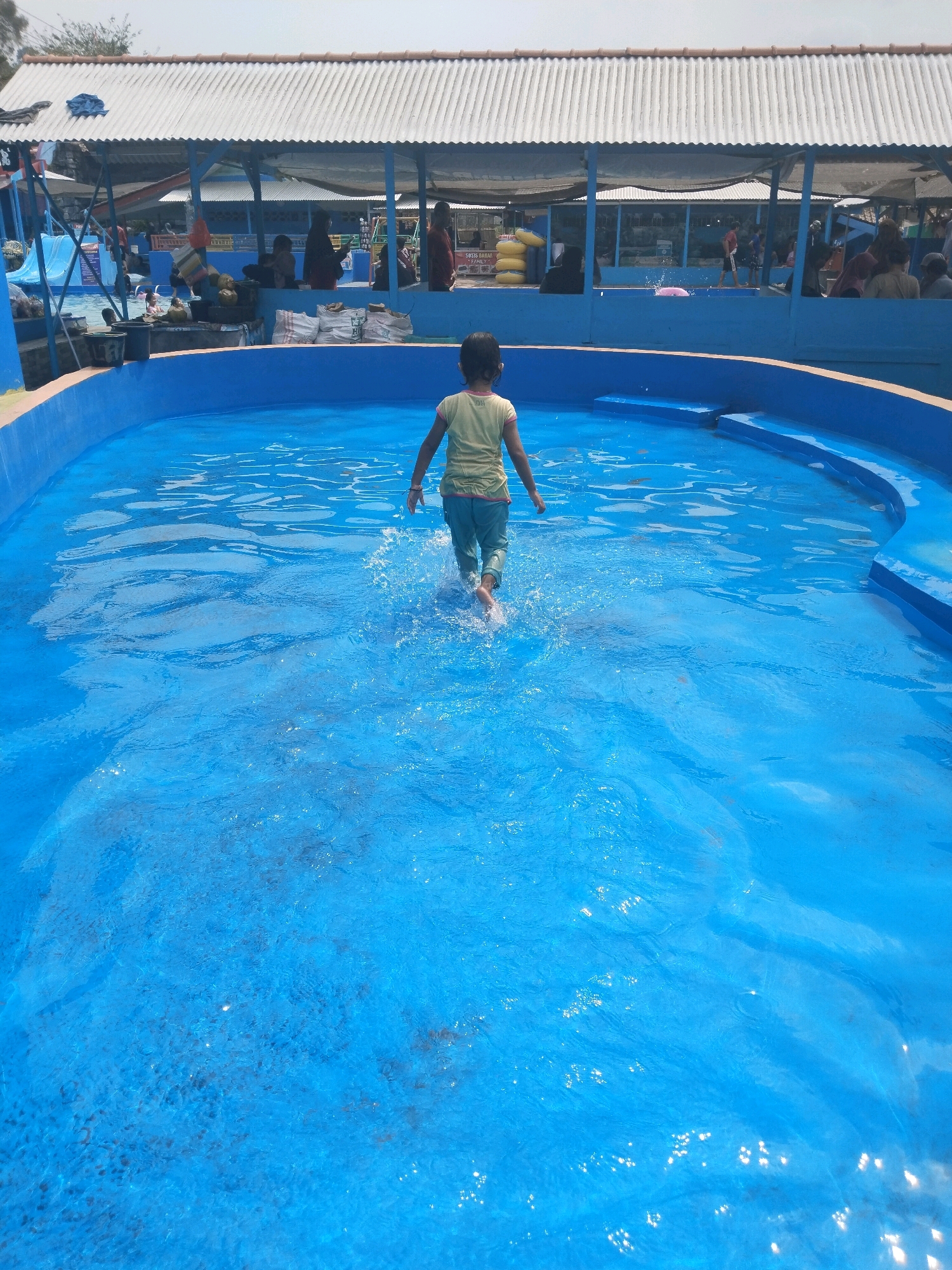 A young girl wearing a green shirt and blue pants walks through a shallow blue swimming pool. She is facing away from the camera and is mid-stride, kicking up water. The pool has a bright blue bottom and sides, with some darker, submerged structures visible. In the background, a covered area with a corrugated metal roof is visible, supported by blue poles. Several people are gathered under this shelter, some sitting and some standing. There are also what appear to be bags of supplies stacked near the shelter. The overall environment suggests an outdoor recreational area, possibly a water park, located in Batukuwung, Indonesia. The lighting indicates it is daytime with bright sunlight.