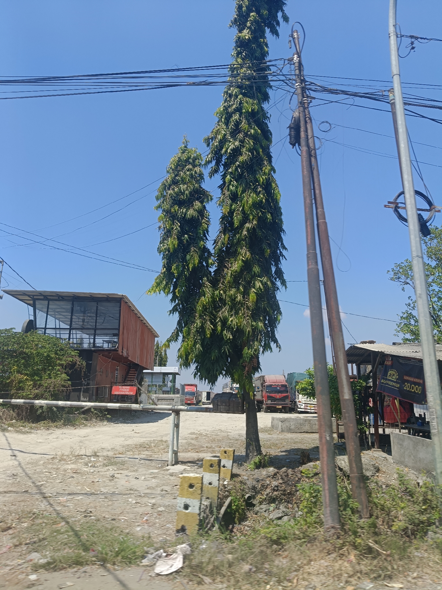 The image depicts an outdoor scene under a clear blue sky, likely in Ambeng Ambeng Watangrejo, Indonesia. In the foreground, a dirt and gravel area is visible with sparse vegetation and scattered debris. A white and yellow striped barrier post is prominent on the right side of the foreground. A white horizontal barrier gate is extended across the scene, suggesting an entrance or checkpoint.
Dominating the midground are two tall, slender evergreen trees, their branches dense with dark green foliage. Behind the trees, a modern-looking building with a cantilevered roof and glass walls is situated on the left. Next to it, several trucks are parked in a dusty lot. On the right side of the midground, a simple open-sided structure with a corrugated roof is present, displaying a banner with text that includes "SUPREMIUM 20.000". Electrical poles and a complex network of overhead wires are visible throughout the image, crossing the sky. The overall impression is of a roadside area with some commercial activity, possibly a logistics or distribution hub, given the presence of trucks and the building's utilitarian design.