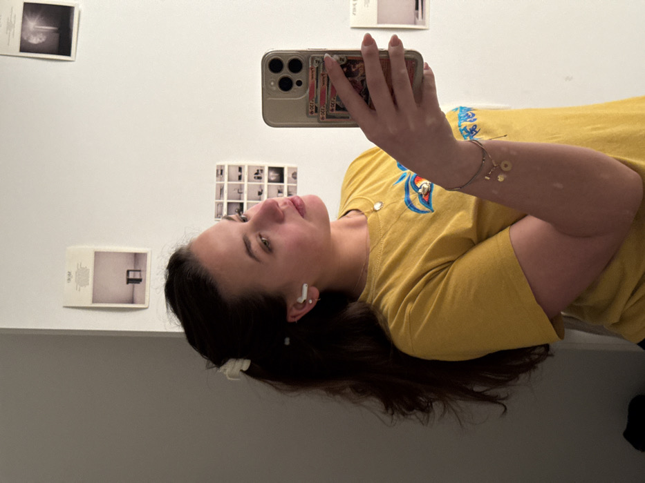 A young woman takes a selfie in a mirror. She wears a yellow T-shirt and wireless earbuds. A phone case featuring Pokémon cards adorns her phone. Her long brown hair is partially clipped up. The background reveals a white wall with small framed pictures or postcards. She has a necklace and bracelets on. The angle is slightly tilted, offering a casual and personal glimpse.
