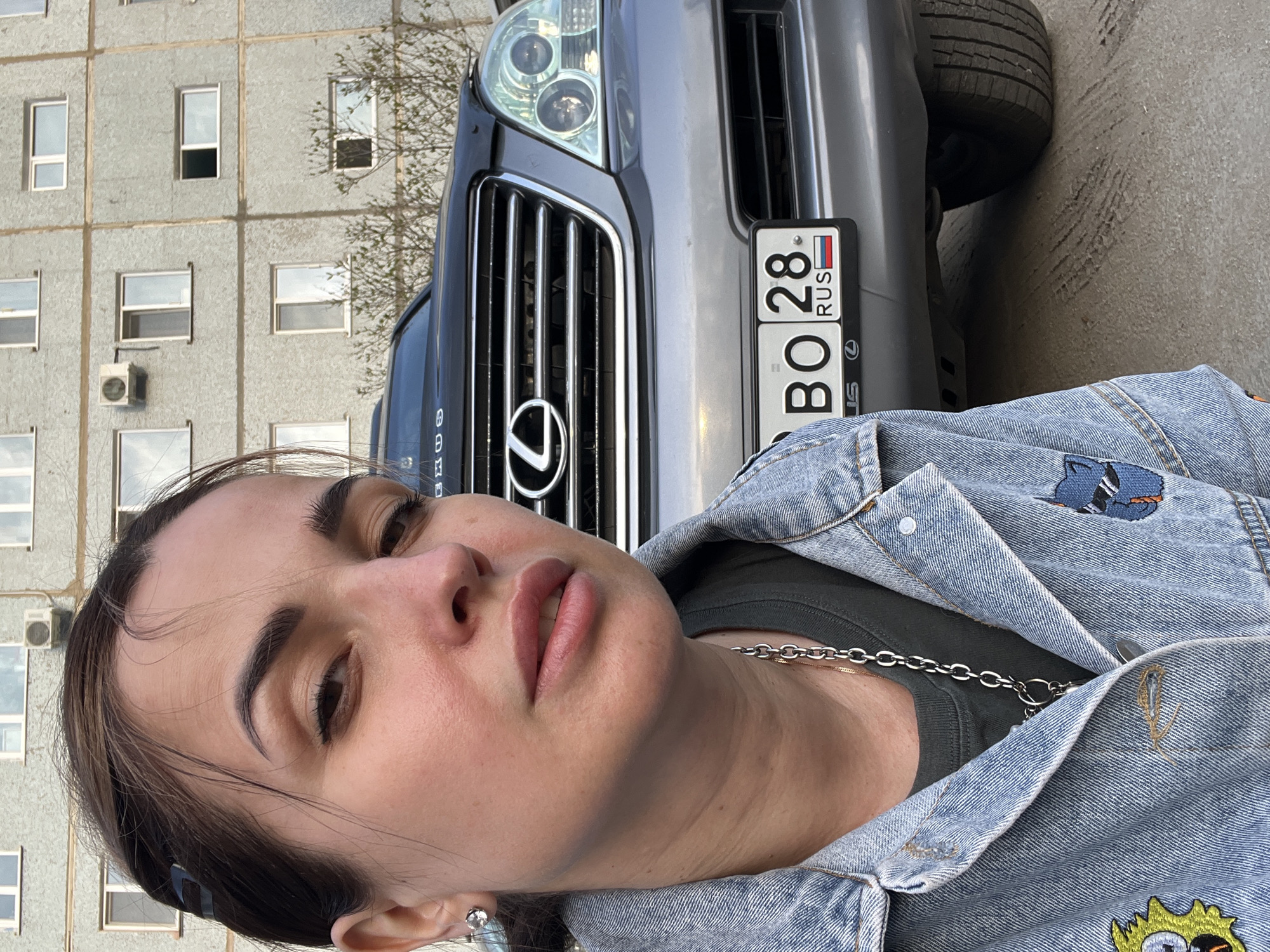 A portrait of a woman in front of a Lexus SUV with a Russian license plate "BO 28 RUS". The background shows a gray concrete building with multiple windows and an air conditioning unit. The location is in an urban area within an unknown city.