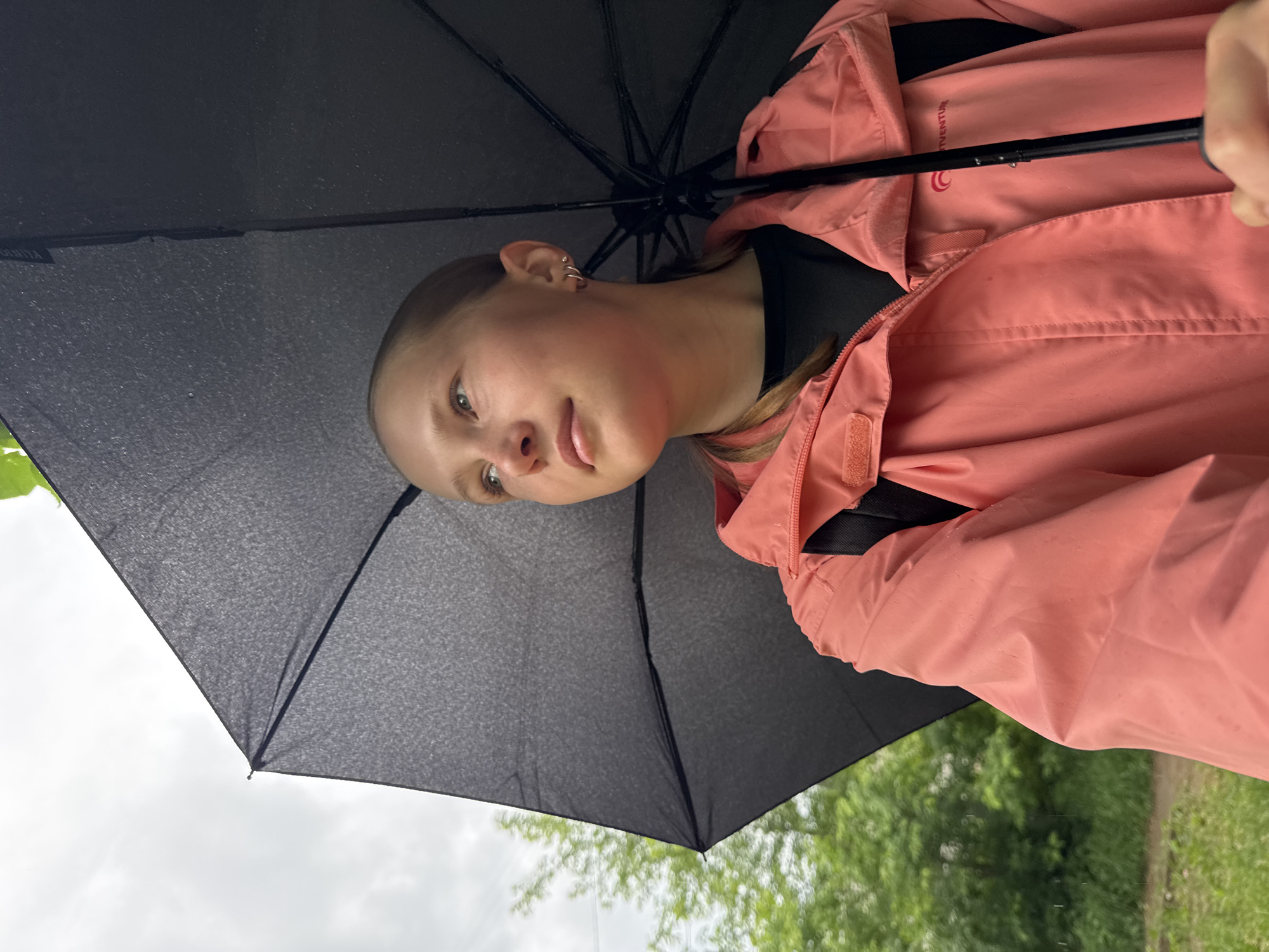 This image features a young person holding a black umbrella outdoors, likely to shield themselves from rain. The subject, who appears to be female with light hair pulled back, is visible from the chest up, looking towards the left side of the frame with a neutral, slightly pensive expression and lightly parted lips. She wears a salmon-pink or peach-colored jacket, which appears to be a waterproof or lightweight outer layer, over a dark shirt. Visible details include small hoop and helix earrings on her left ear.
The large black umbrella is held open, its underside occupying a significant portion of the upper left side of the frame, showing its ribs and a textured fabric. Part of the umbrella's shaft is visible, held by the person's hand. On the umbrella's fabric, and more clearly on an inner flap of her jacket, the text "ADVENTUR" (likely part of "ADVENTURE") is visible, alongside a circular logo resembling a stylized 'C' or spiral.
The background suggests an overcast day in a natural outdoor setting, indicative of Chaikovsky, Russia. A cloudy, grey sky is visible beyond the umbrella, while blurred green foliage, including trees and grass, fills the right and lower-right parts of the frame, hinting at a park or a path amidst nature. The overall scene captures a quiet moment during an outdoor activity under potentially damp weather conditions.