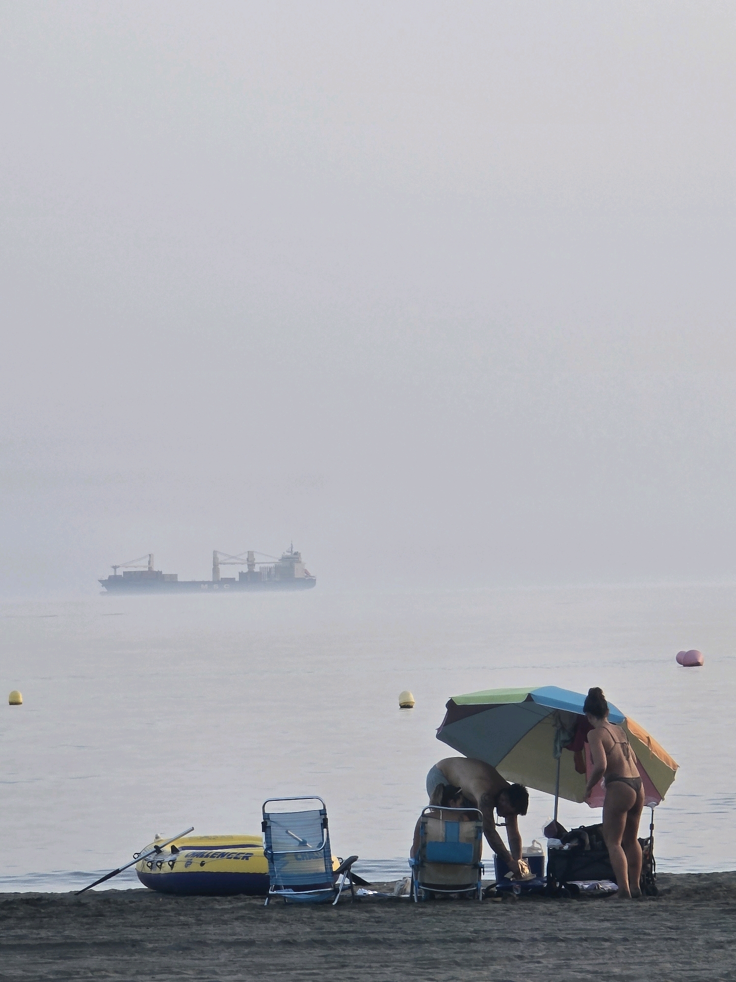 The image depicts a hazy beach scene in Málaga, Spain, with a cargo ship visible in the distance on a calm sea. In the foreground, two people are preparing for a day at the beach under a multi-colored umbrella. A man, wearing light-colored shorts, is bent over a cooler and appears to be retrieving something. A woman, wearing a dark bikini, stands next to him, facing away from the camera. They are surrounded by beach gear, including chairs and bags. To the left, a yellow and blue inflatable boat with oars is resting on the dark sand, with a blue beach chair nearby. Several buoys are scattered in the shallow water. The overall atmosphere is serene, with the soft light and gentle waves contributing to a peaceful ambiance.