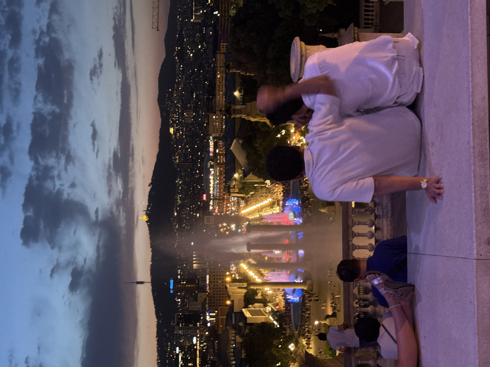 This is an outdoor, ground-level, medium shot taken at dusk, looking over a cityscape. The foreground features four individuals seated on a stone ledge. Two men, facing away from the camera, are embracing. One man has his arm around the other, and both are wearing white shirts. To their left, a person in a white top and dark pants is visible from the waist down, with their legs crossed. Further left, another individual in a light-colored shirt and dark pants stands facing the cityscape.
In the mid-ground, a large, illuminated fountain is the central focus, with water jets shooting upwards and colored lights reflecting off the water. The fountain is situated in a plaza filled with people, and beyond that, rows of buildings with illuminated windows stretch into the distance. The cityscape is nestled against a hill.
In the background, the sky is a blend of deep blue and dark gray clouds, with a hint of orange near the horizon. A tall communication tower stands on the silhouette of the hill, and lights from the city twinkle below. The overall scene suggests a view from a vantage point overlooking the Magic Fountain of Montjuïc in Barcelona, Spain, during an evening performance.