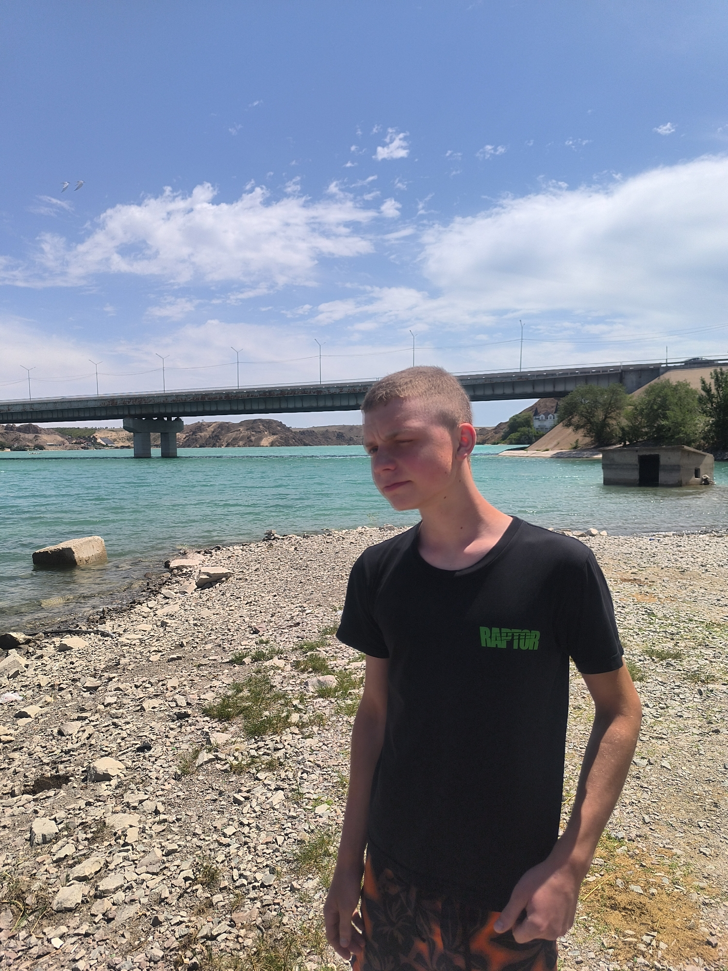The image shows a young boy standing on a rocky shoreline with a turquoise body of water behind him. In the background, a large bridge spans the water, with a small, submerged concrete structure to the right of the frame. The sky is blue with scattered white clouds, and two birds are visible in the upper left corner. The boy is wearing a black t-shirt with a green "RAPTOR" logo and patterned shorts. The environment appears to be a reservoir or large lake, consistent with the Kapchagay Reservoir located near Qonaev, Kazakhstan, which is known for its distinctive turquoise waters and the presence of bridges and structures along its banks. The lighting suggests it is daytime, likely sunny given the shadows.