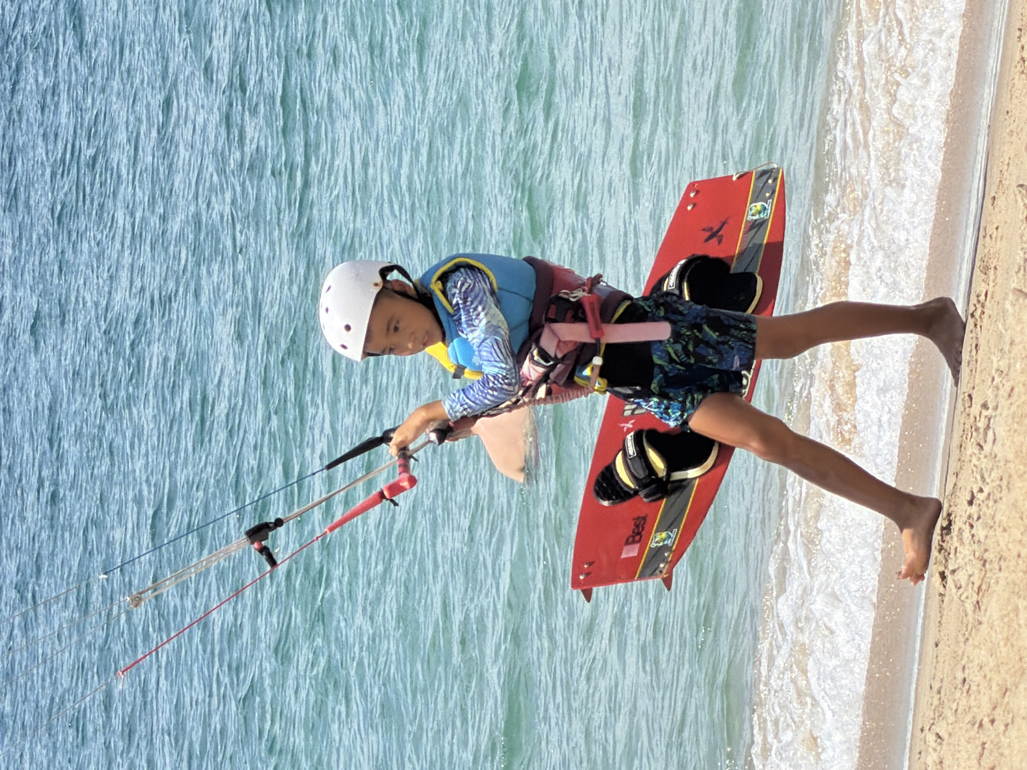 A young boy, wearing a white helmet and a blue and yellow life vest, walks along the sandy shore of Nha Trang, Vietnam, carrying a red kiteboard. He is holding onto the control bar of a kite, which is out of frame. The water is a clear turquoise with small waves breaking on the beach. In the background, another person is partially submerged in the water. The boy is wearing patterned swim trunks and appears to be preparing to kite surf.