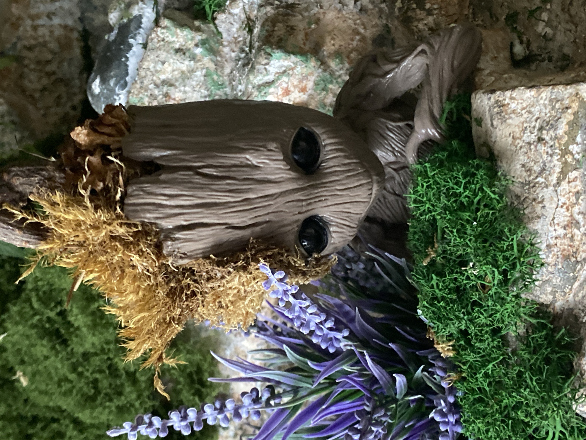 A small, detailed diorama featuring a figure resembling the character Groot from Guardians of the Galaxy is the central subject. The figure is brown and textured to resemble wood, with black eyes. It is placed within a miniature landscape. To the left of the figure are artificial lavender sprigs with small purple flowers and slender green leaves. In front and to the left of the figure is a clump of bright green, textured artificial moss. Behind the figure, the diorama incorporates faux rocks and green mossy textures. The lighting suggests an indoor setting. The scene does not contain any identifiable elements that would allow for its location to be determined, including in the city of Novokuznetsk, Russia.