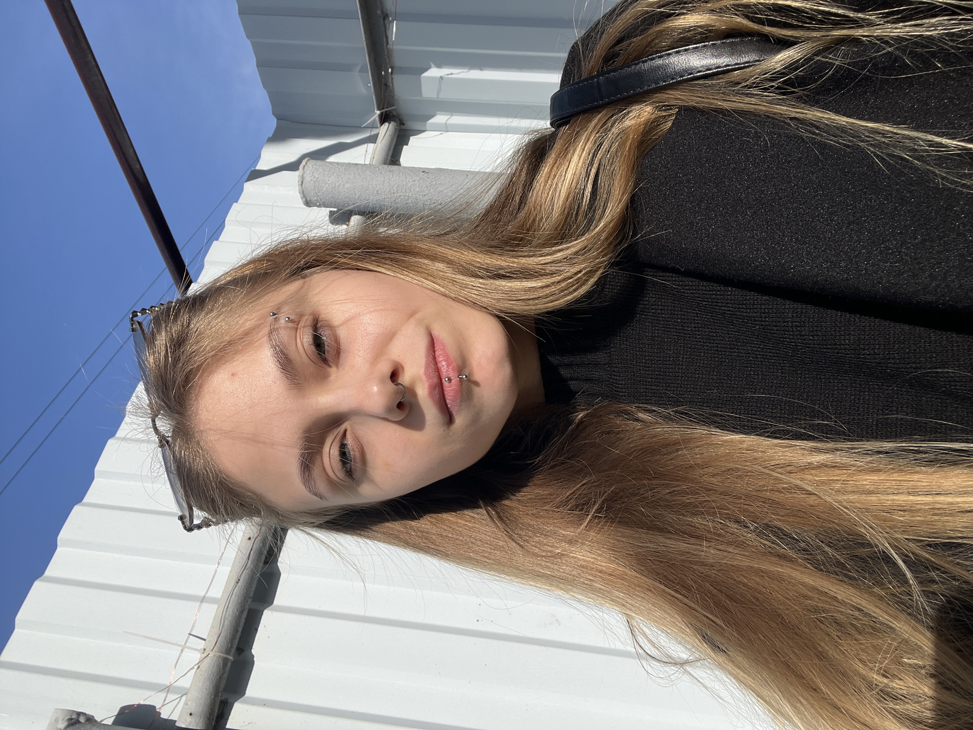 A young woman with blonde hair is lying outdoors in bright sunlight. She has piercings in her eyebrow, nose, and lip. She is wearing a black sweater. Behind her are sections of white metal roofing. A metal pole runs behind her and out of frame. The background includes a clear blue sky.
Based on the user query, the location is assumed to be in the city of Фоки, Russia.