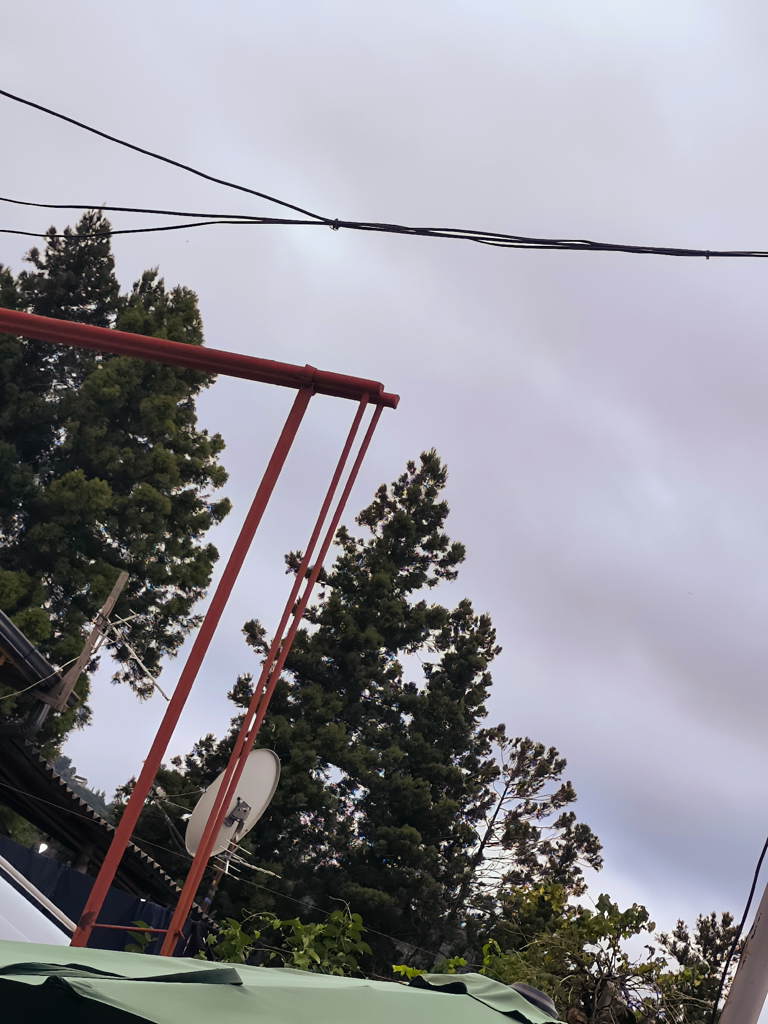 A low-angle outdoor shot captures a cloudy sky, bisected by power lines in the upper portion. A red metal structure, likely part of a swing set or similar apparatus, dominates the left and central areas of the frame. Trees with dark green foliage are visible behind the red structure, their branches reaching towards the sky. A white satellite dish and an antenna are mounted on a pole behind the red structure. In the foreground, a section of a green tarp or awning is visible at the bottom of the image. The overall impression is that of an urban or suburban environment, with a mix of man-made structures and natural elements.